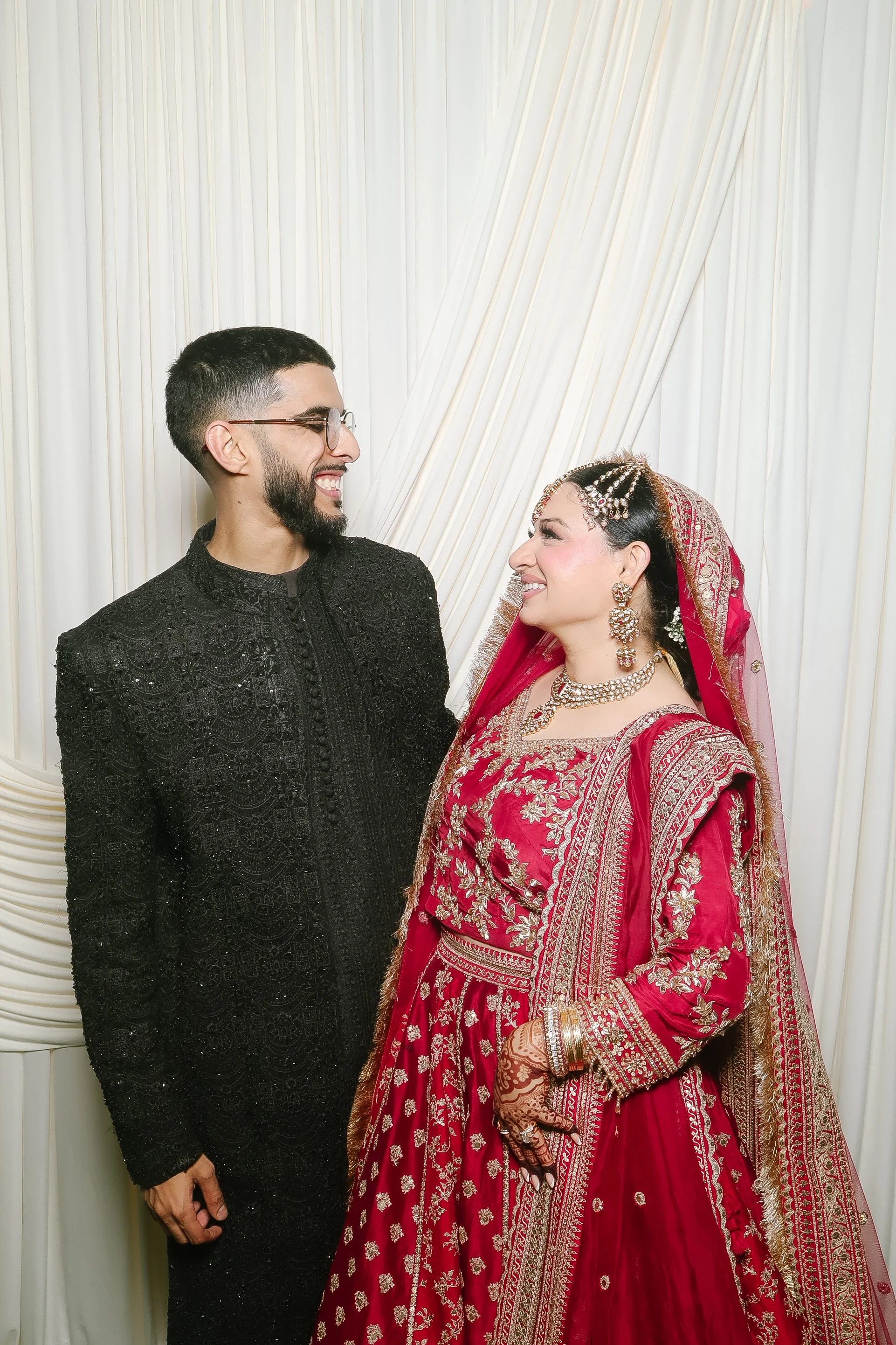 Wedding couple in traditional South Asian attire smiling at each other in front of Bella Photobooth’s ivory drape backdrop.