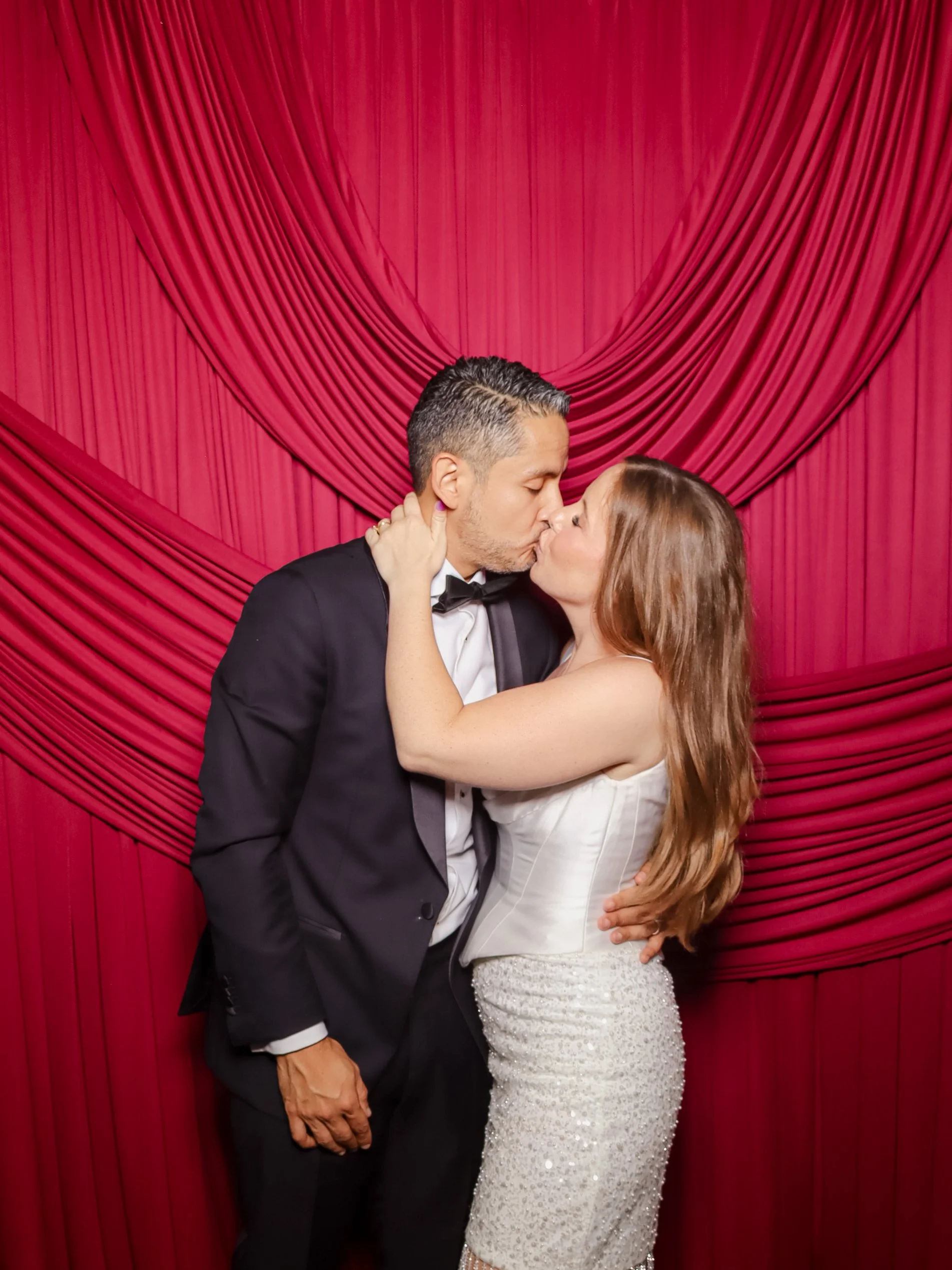 Couple with red drape
“Bride and groom sharing a kiss at their wedding photo booth with an elegant red drape backdrop.”