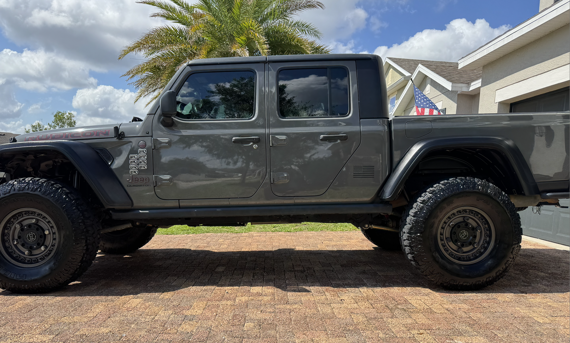 A gray Jeep Rubicon Gladiator pickup truck parked in a driveway with a brick surface in front of a house. The background includes a large palm tree, partly cloudy sky, and the house has beige siding, a gray roof, and an American flag hanging near the