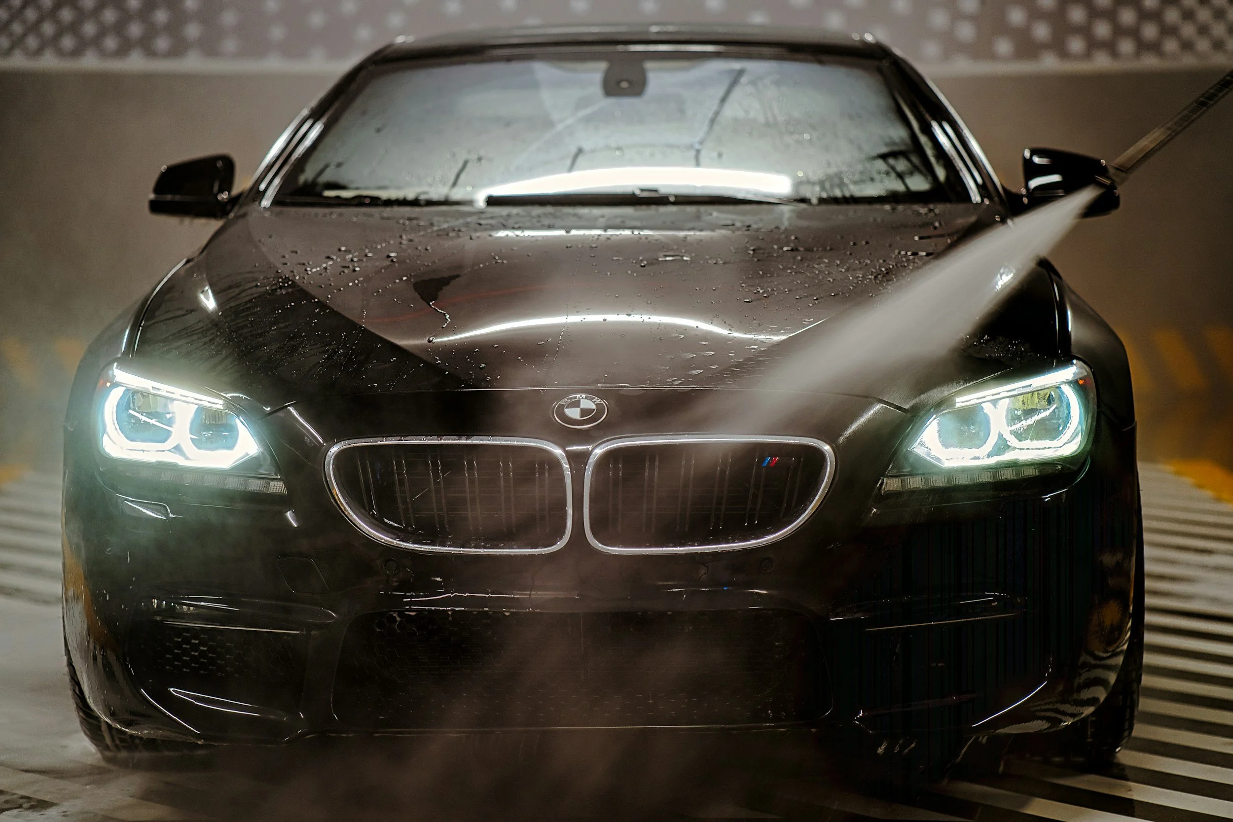 A close-up of a black BMW car with water droplets on its hood, headlights turned on, in an indoor parking garage.