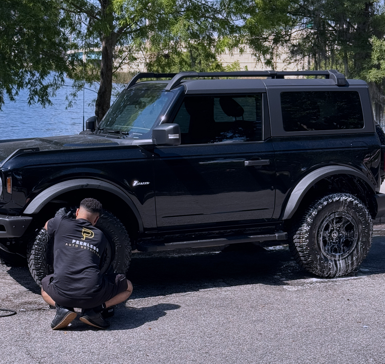 Person cleaning the wheel of a black SUV parked outdoors near trees, by a lake or river.