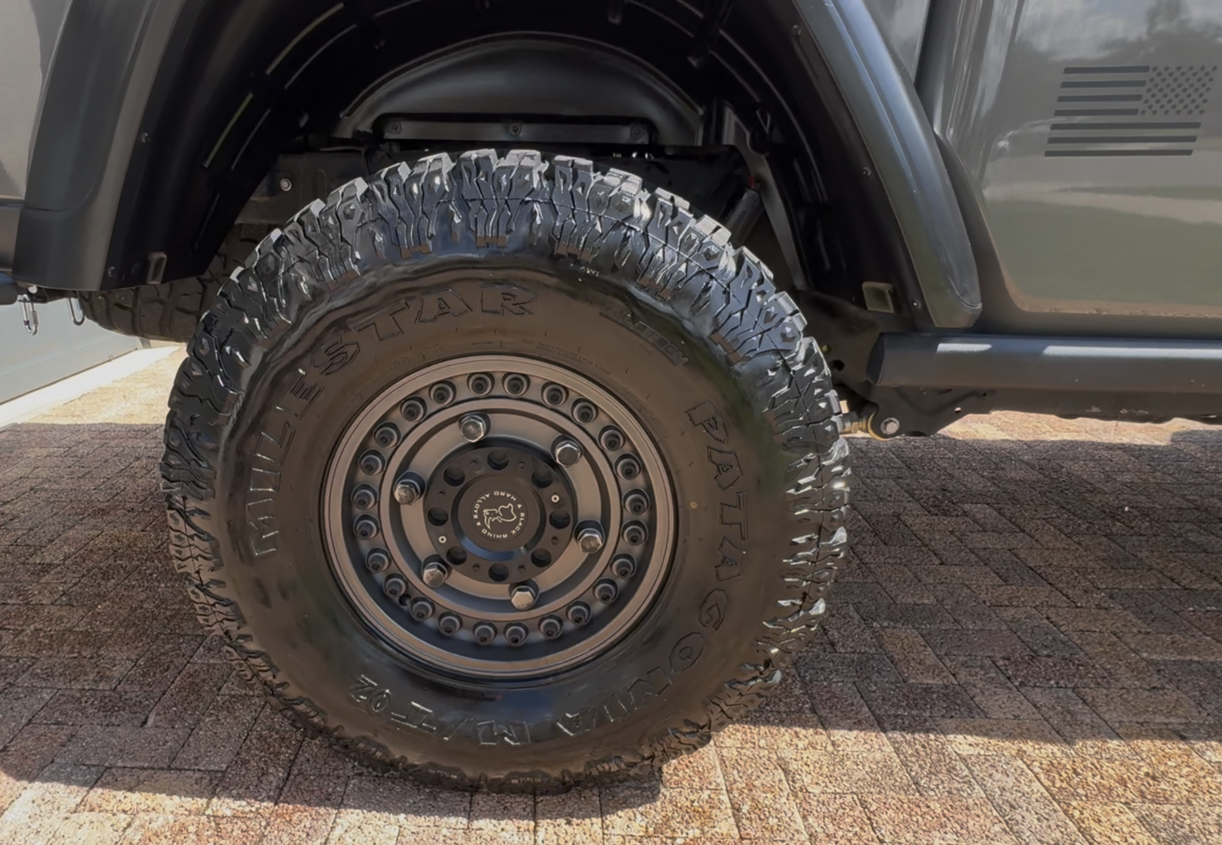 Close-up of a large off-road tire with deep tread pattern mounted on a vehicle, with part of the wheel well and vehicle body visible.