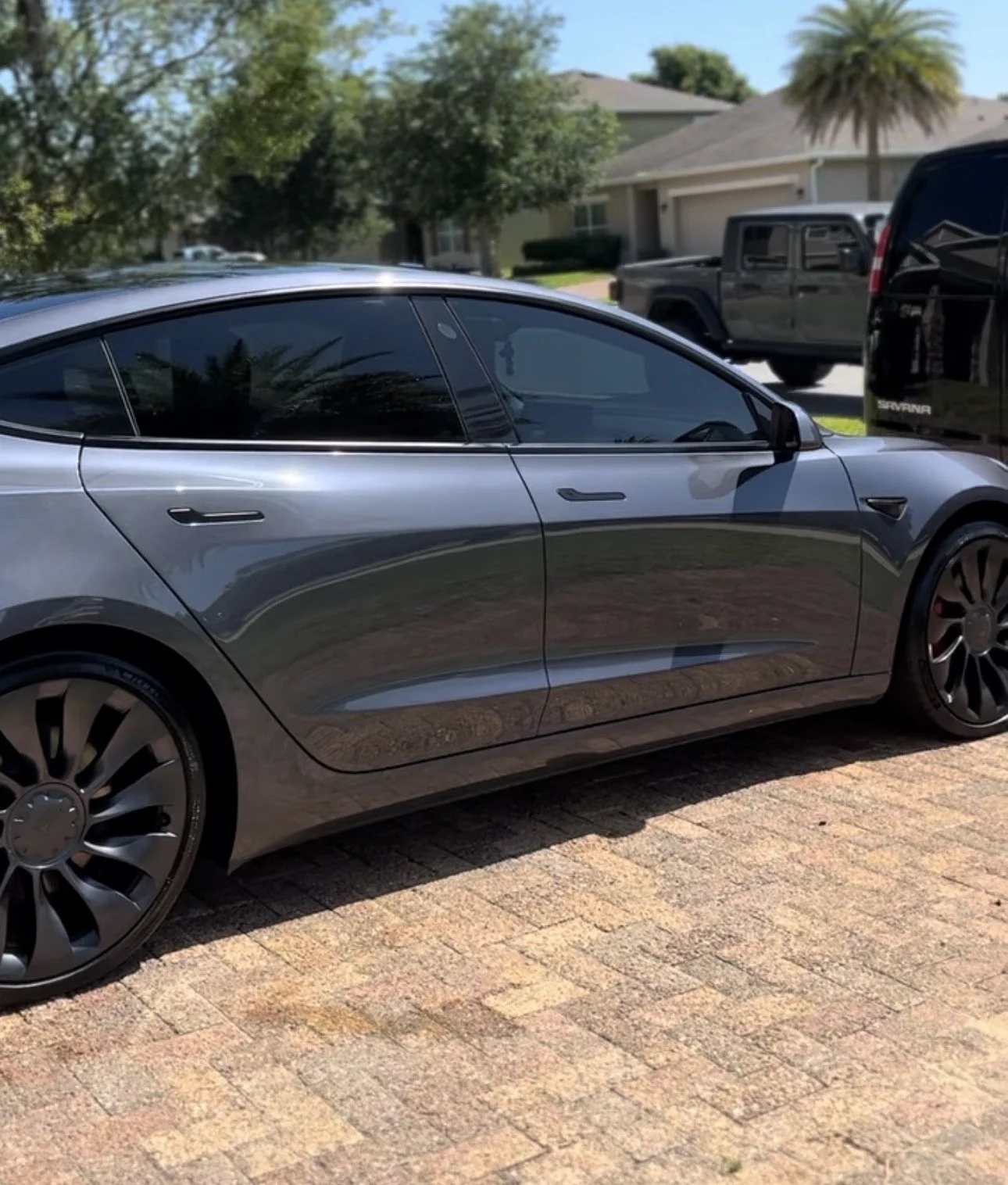 A silver Tesla Model 3 parked on a brick driveway in front of a suburban house with trees and other vehicles in the background.