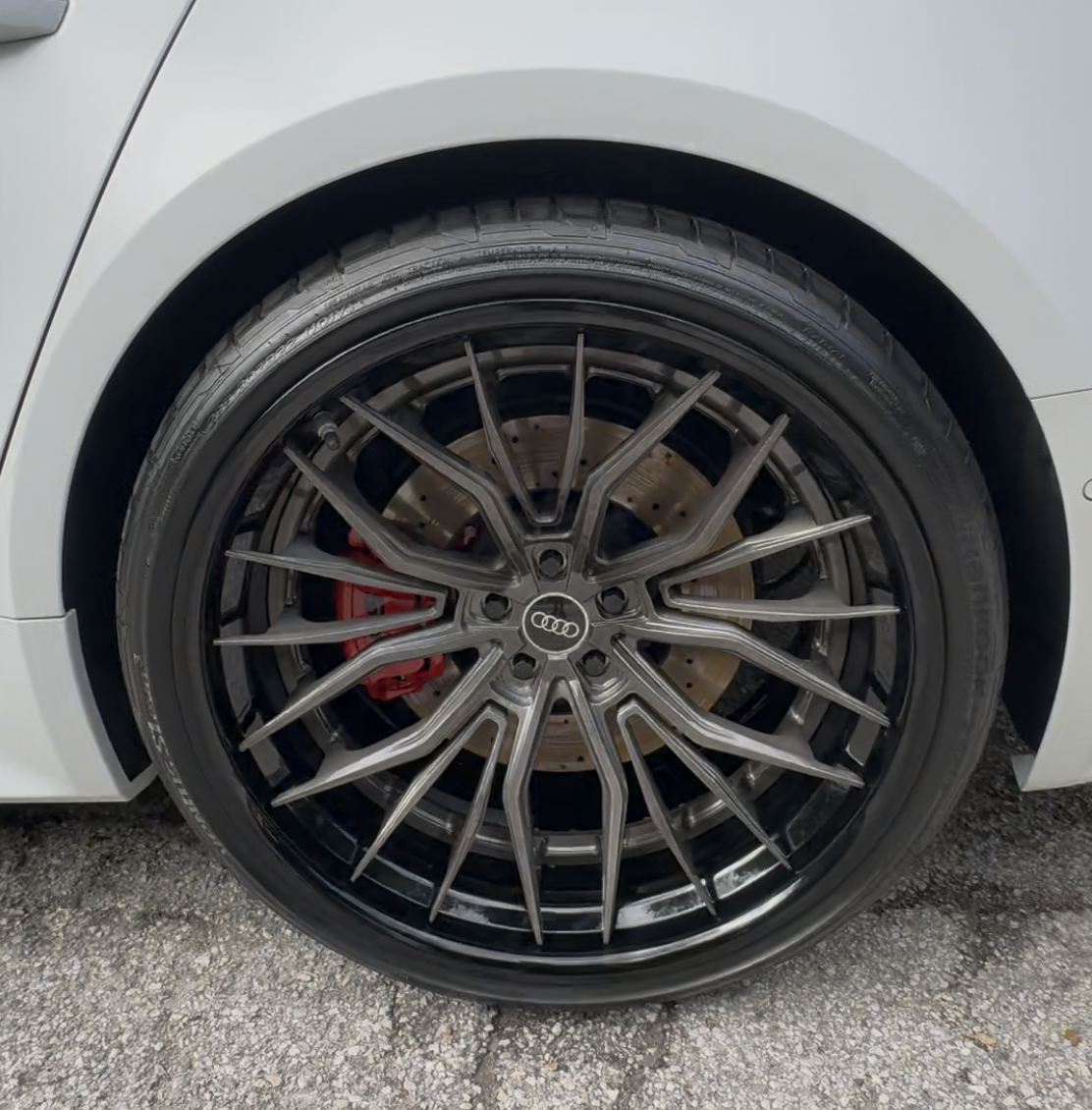 Close-up of a car wheel with a black alloy rim featuring intricate spoke designs, a visible brake disc and red brake caliper, on a textured asphalt surface.