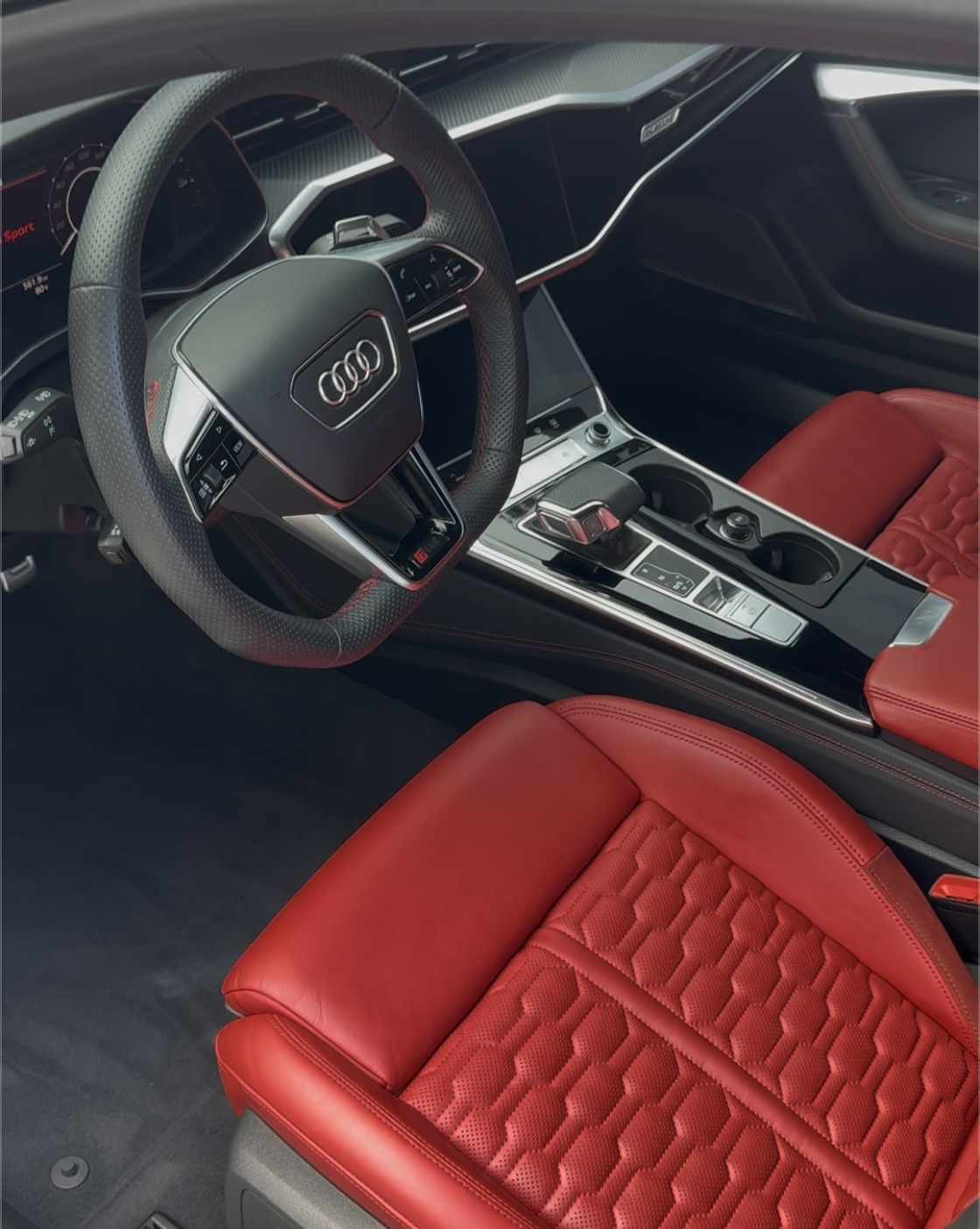 The interior of an Audi sports car featuring a black steering wheel with the Audi logo, a digital dashboard, a center console with gear controls, and red leather seats with quilted stitching.