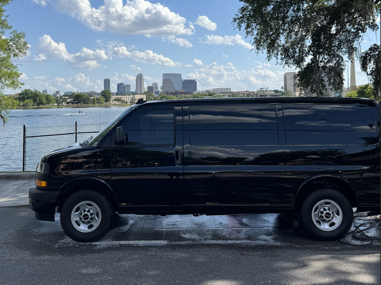 Black van parked near water with city skyline in the background, trees on the sides, and a partly cloudy sky.