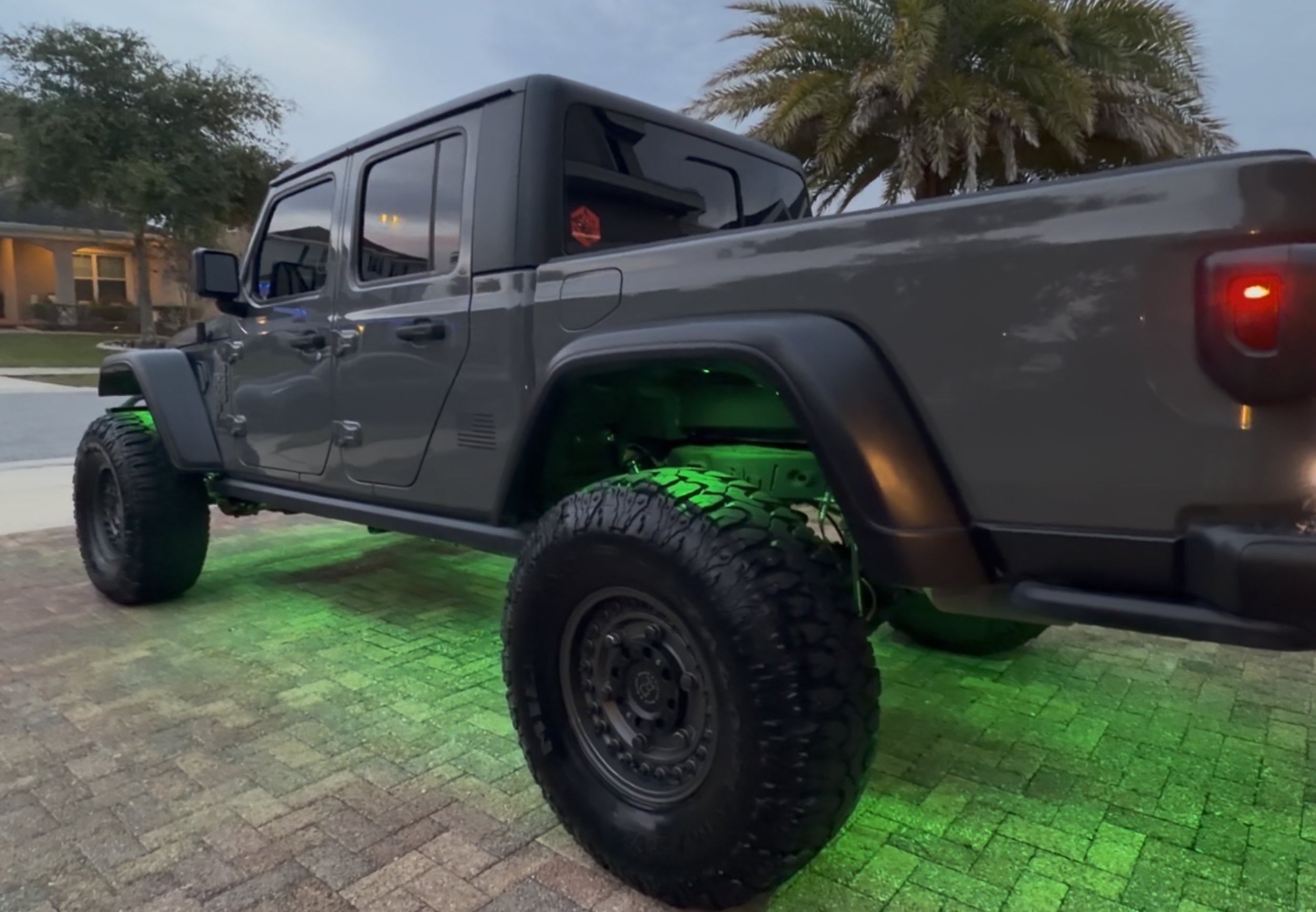 A gray Jeep Gladiator pickup truck parked on a brick driveway at dusk, illuminated by green underglow lights, with trees and a house in the background.