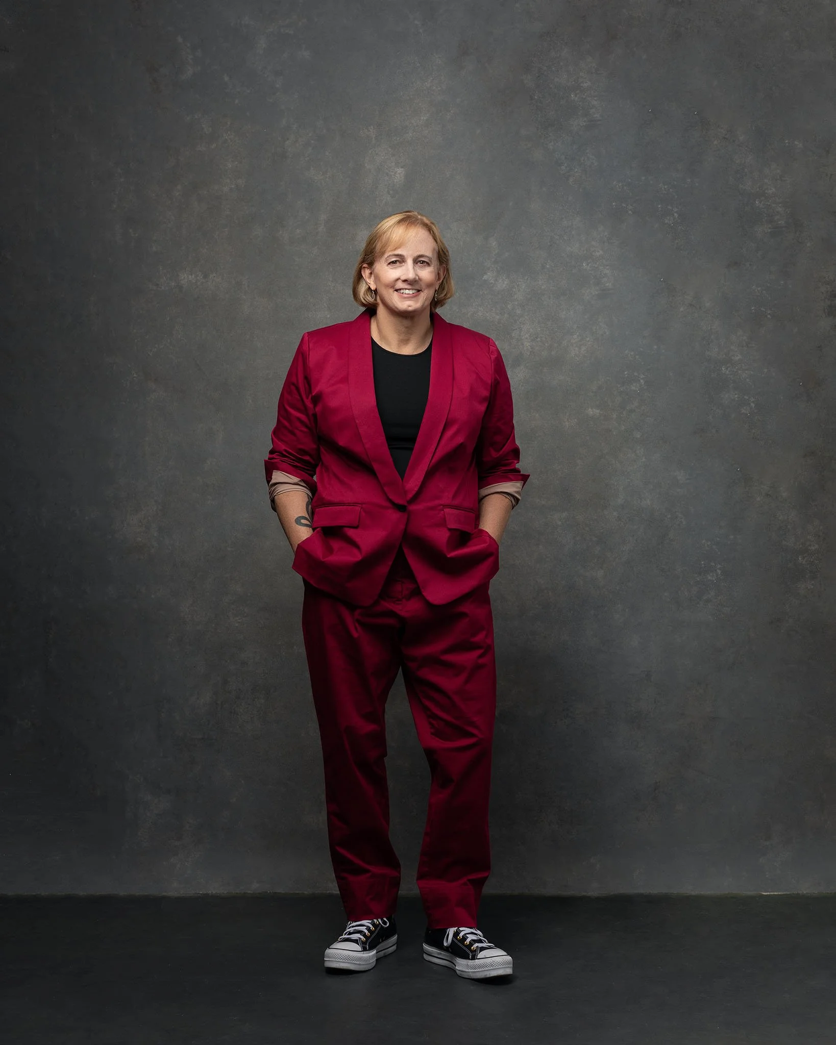 Jess is wearing a dark red suit coat and matching trousers. She wears a black t-shirt under the coat. She is standing with her hands in her pockets, sleeves pulled back. She is wearing black custom Converse high-top Chucks. She is smiling.