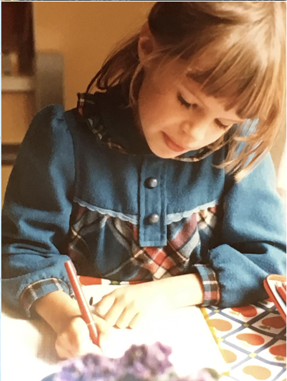A young girl with brown hair and bangs, wearing a denim and plaid jacket, is drawing with a red marker at a table with colorful patterned paper.