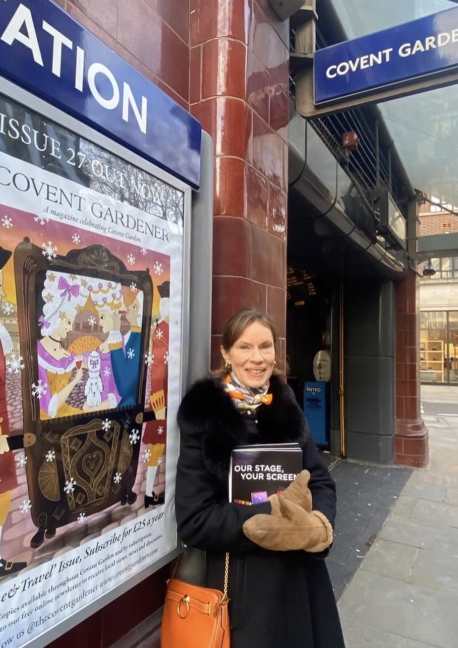 A woman standing on a city sidewalk outside a metro station, holding a brochure, smiling, with a decorated poster for Covent Gardener nearby.