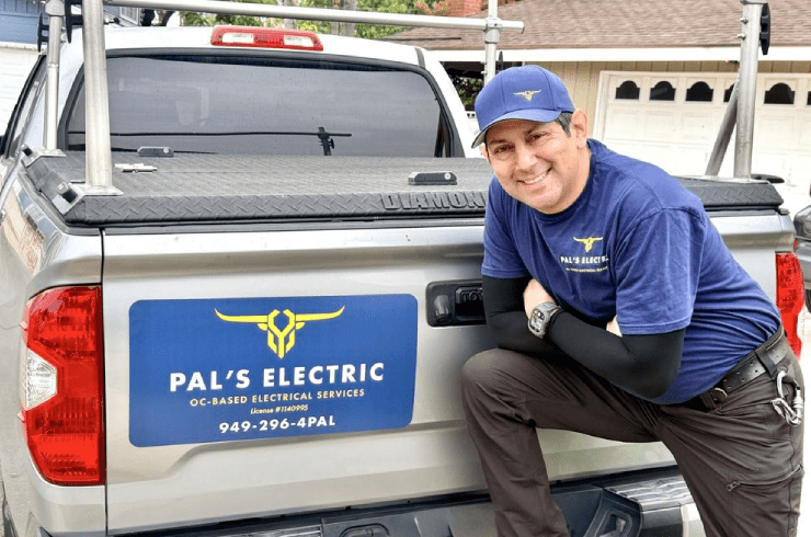 A smiling man in a blue Pal's Electric T-shirt and cap, with black arm sleeves, posing next to a pickup truck with a Pal's Electric sign, outdoors in a residential area.