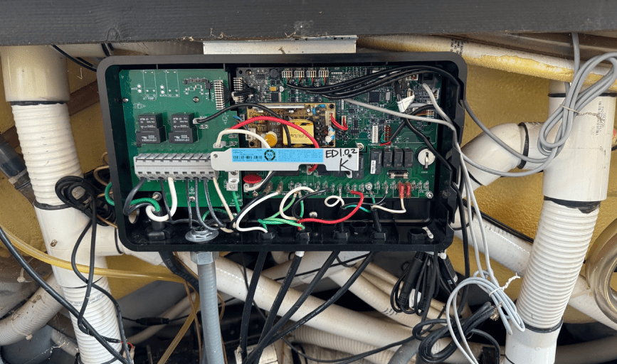 Open electrical control box with circuit boards, wires, and electronic components inside, mounted on pipes and behind a structure.