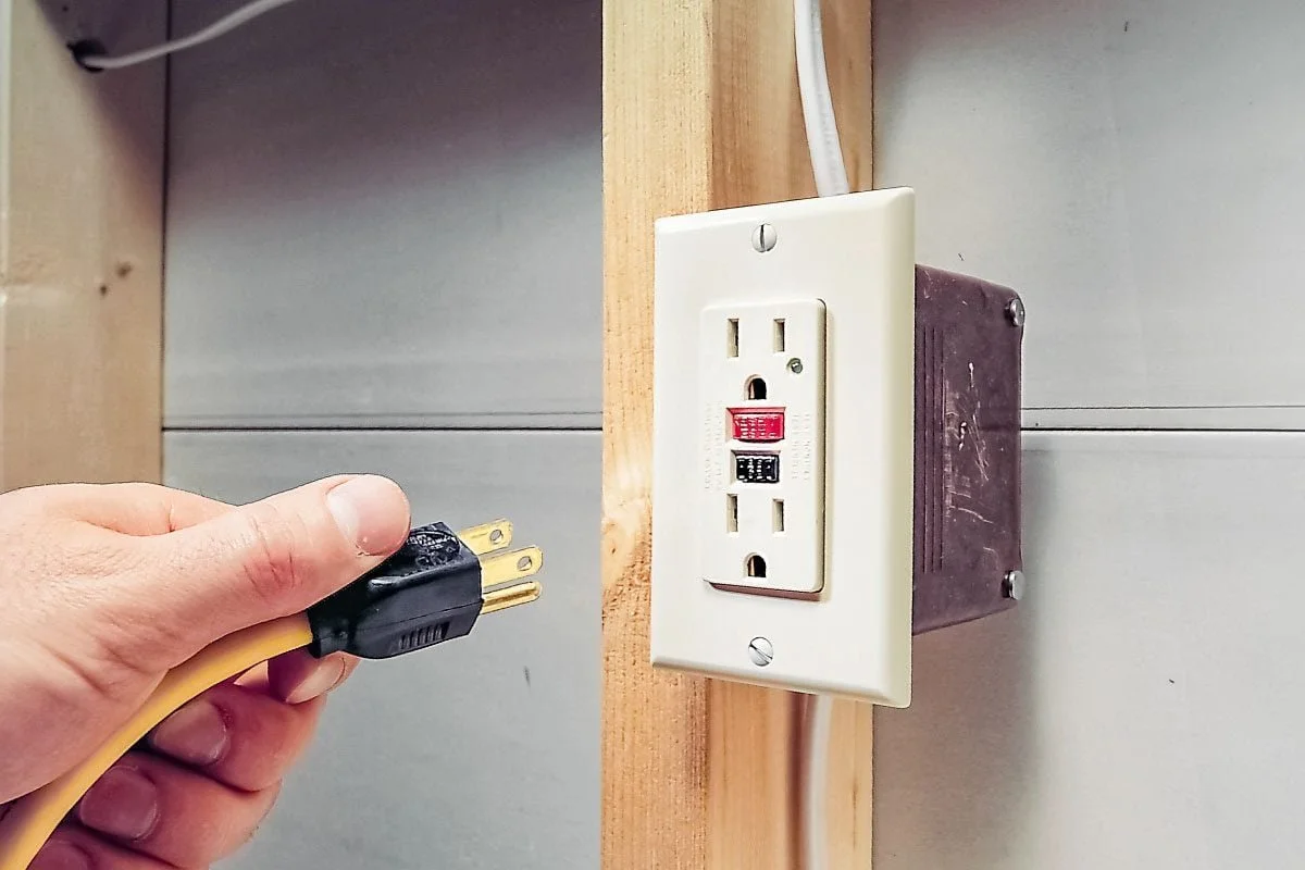 A person holding a yellow electrical cord near an electrical outlet with a built-in circuit breaker switch.
