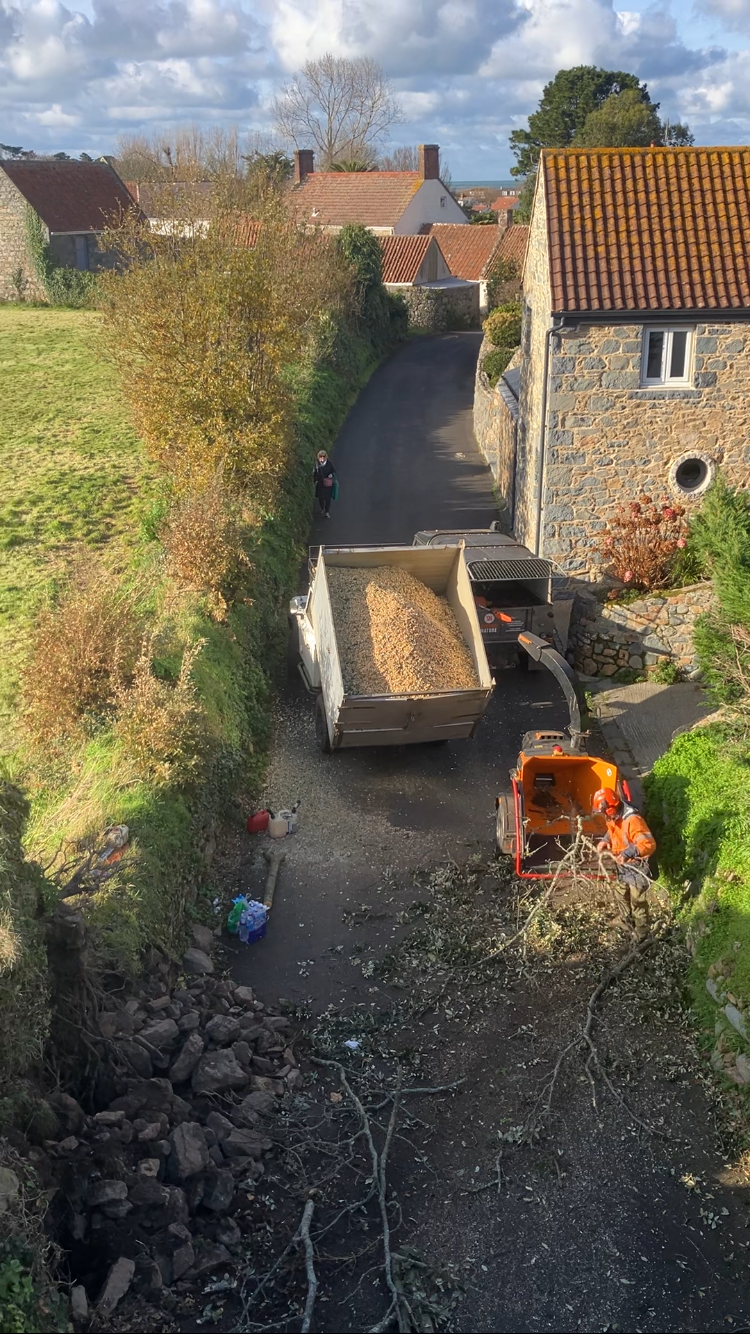 Workers using machinery to remove tree branches and debris from a small driveway next to residential houses on a rural street, with a truck filled with wood chips and a person walking along the road.