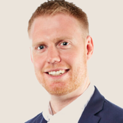 A smiling man with red hair and a beard wearing a dark suit and tie on a light background.