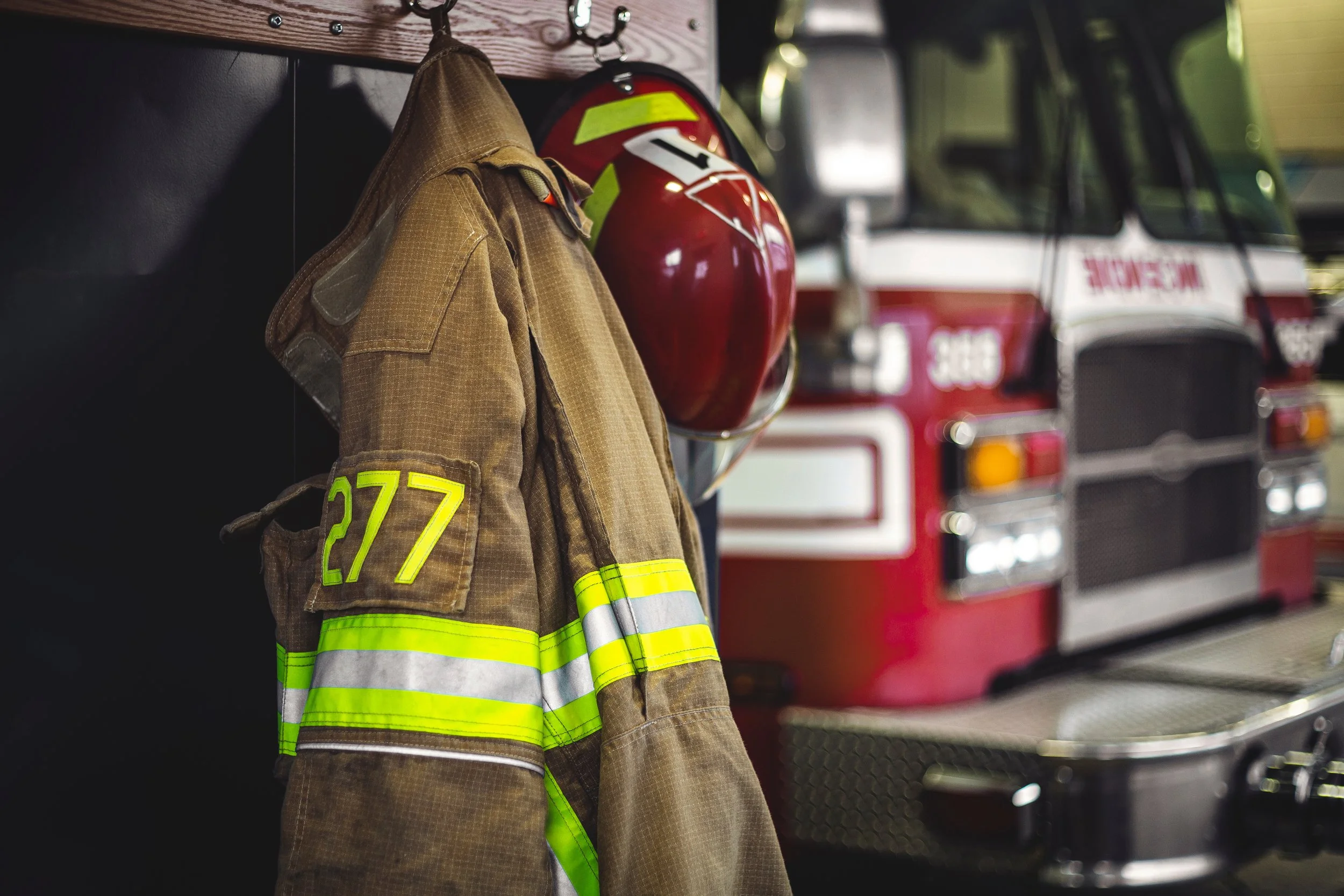 Firefighter's jacket hanging on a hook with a red firefighter helmet nearby, with a fire truck in the background.