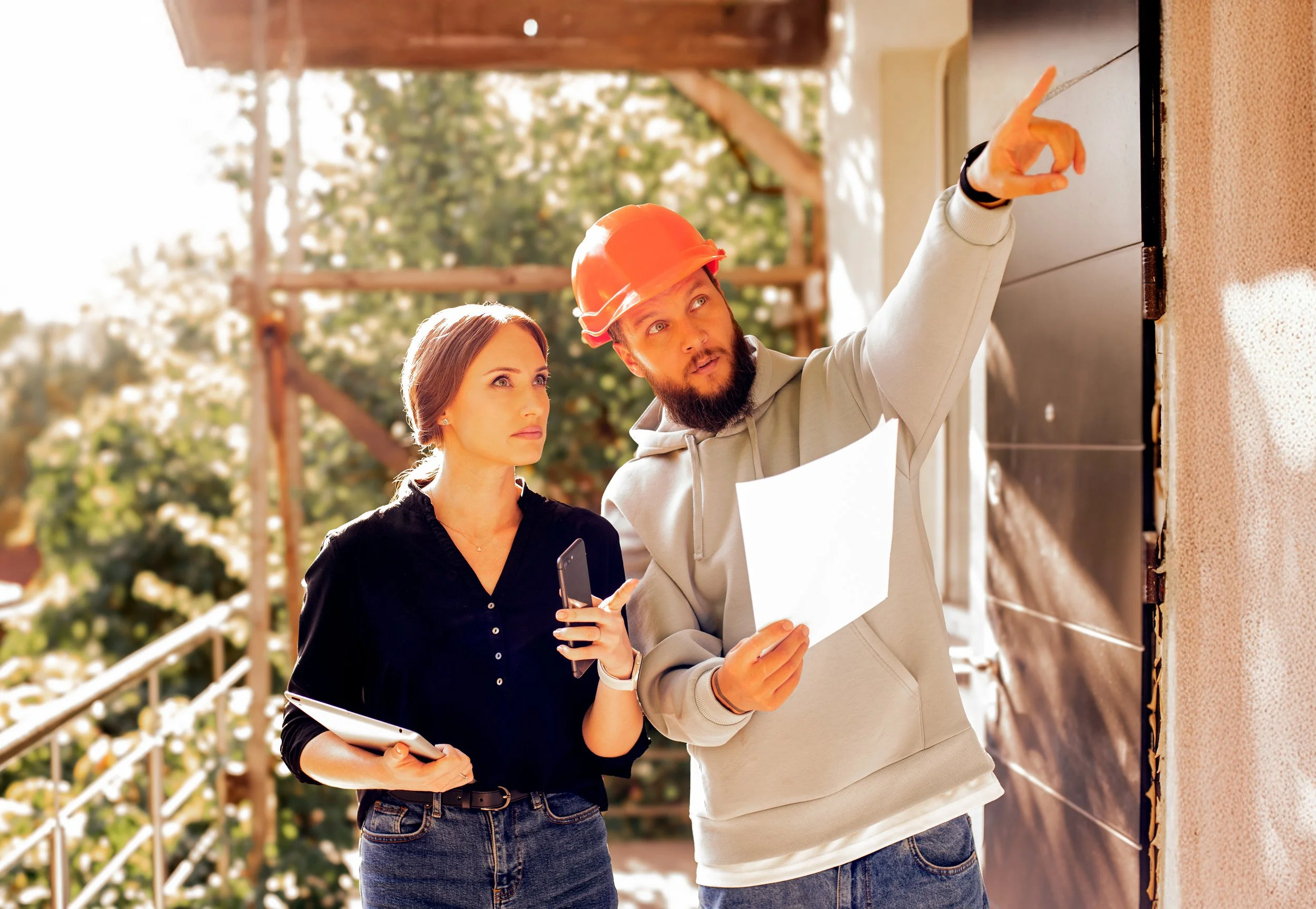 A man in a gray hoodie and an orange construction helmet explains something to a woman holding a folder and a phone, standing outside near a building under construction with the outdoors and trees in the background.