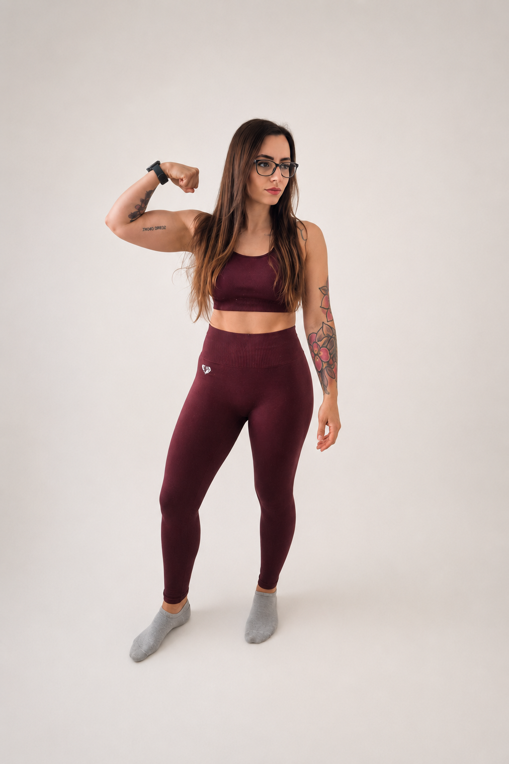 A woman with long brown hair, glasses, and tattoos on her arms, wearing a matching maroon workout crop top and leggings, gray socks, flexes her right arm to display her bicep in front of a plain white background.