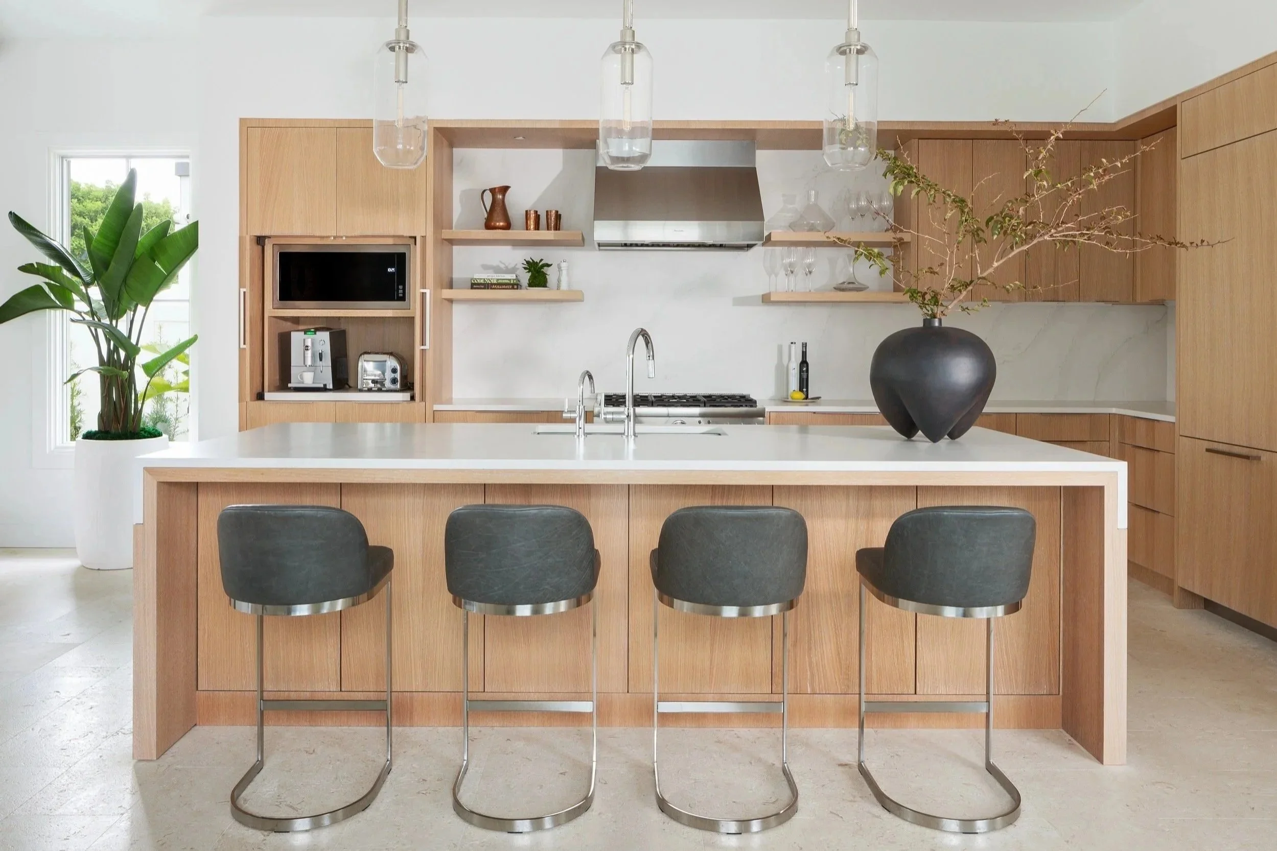 A kitchen with white countertops, light wood cabinets, glass pendant lights, and a stainless steel hood.