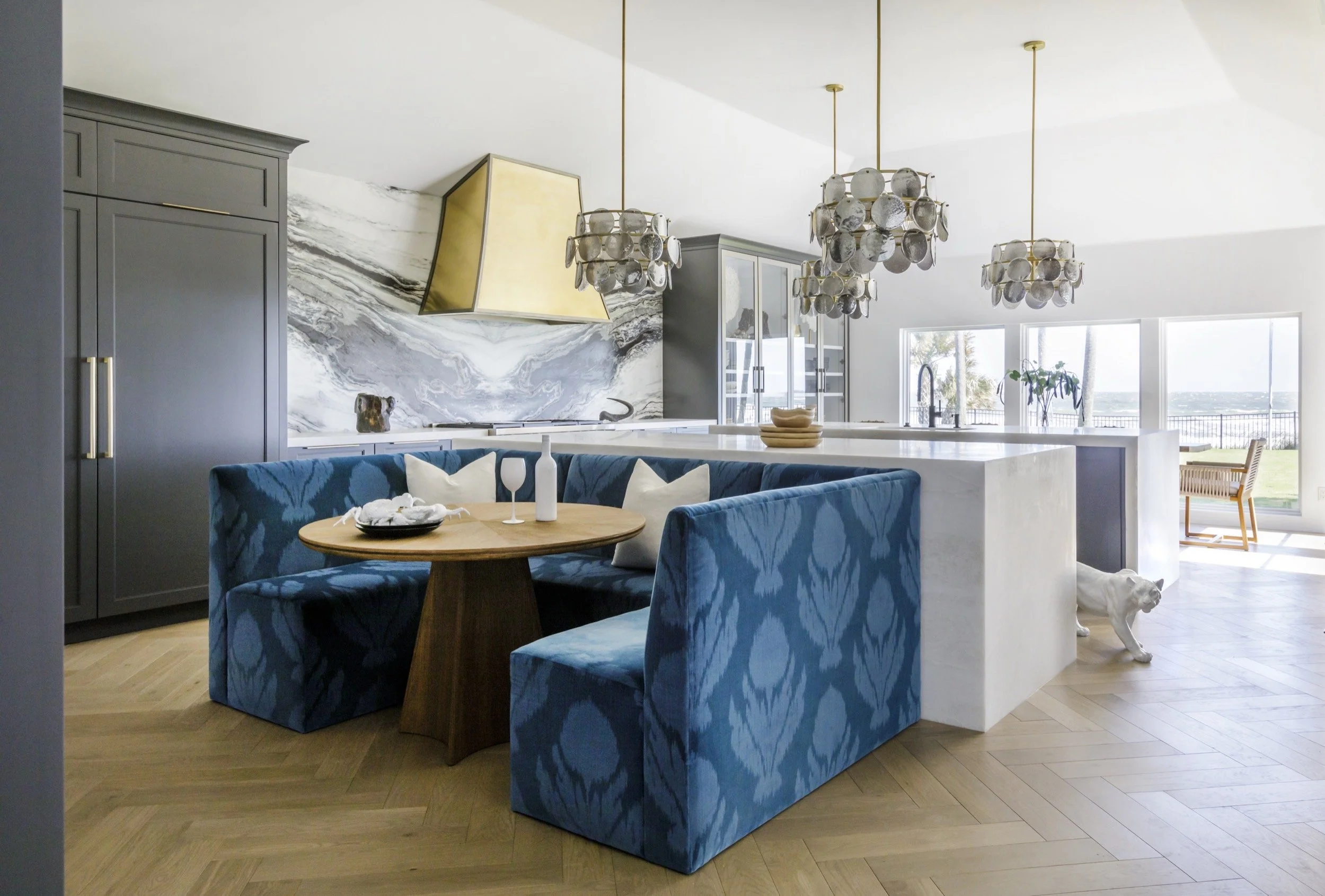 A large kitchen with two white kitchen islands, grey cabinets, a bold marble backsplash, a gold hood, and a blue dining nook.