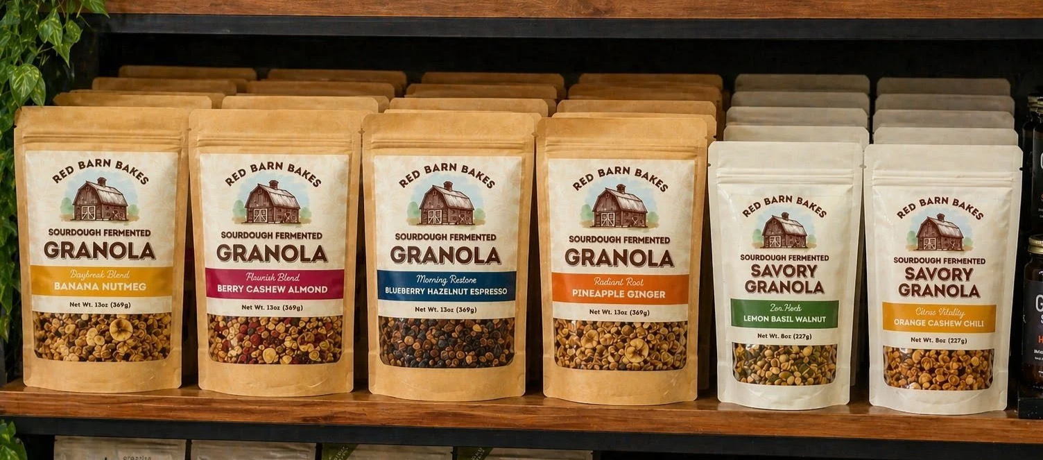 Shelf displaying six different varieties of Red Barn Bakes sourdough fermented granola in resealable pouches with labels, including Banana Nutmeg, Berry Cashew Almond, Blueberry Hazelnut Espresso, Pineapple Ginger, Lemon Basil Walnut, and Orange Cashew Chili.