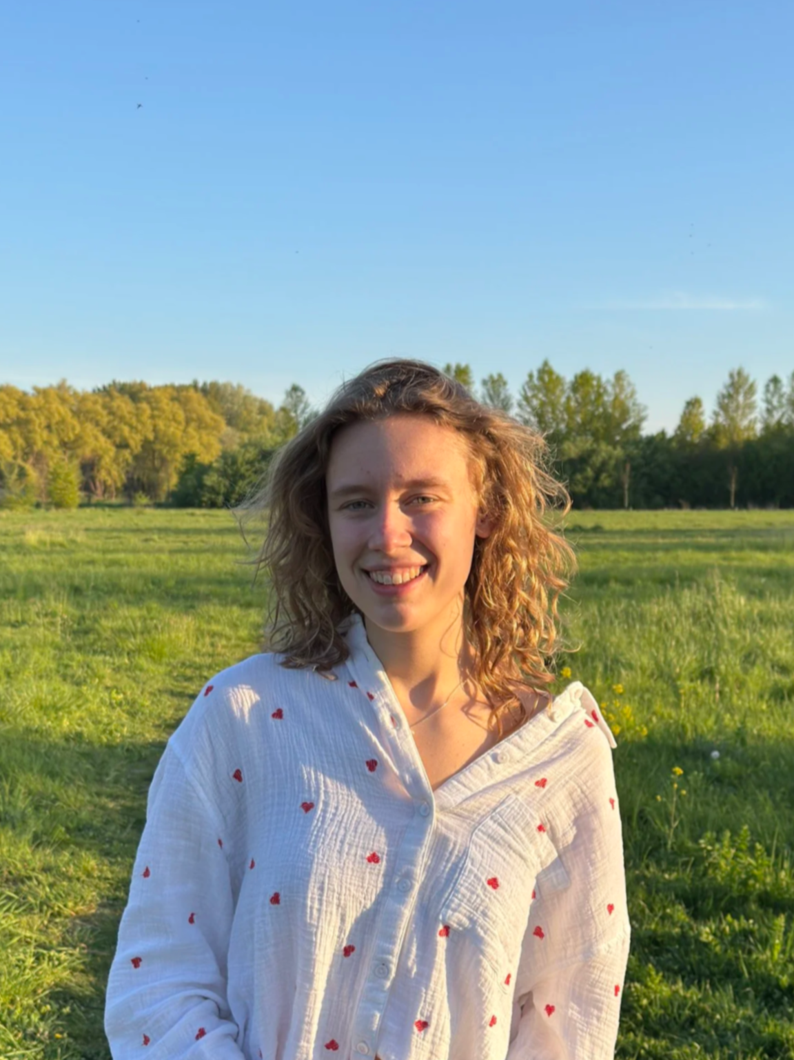 Een meisje met golvend haar in een veld, glimlachend, met bomen op de achtergrond en een blauwe hemel.