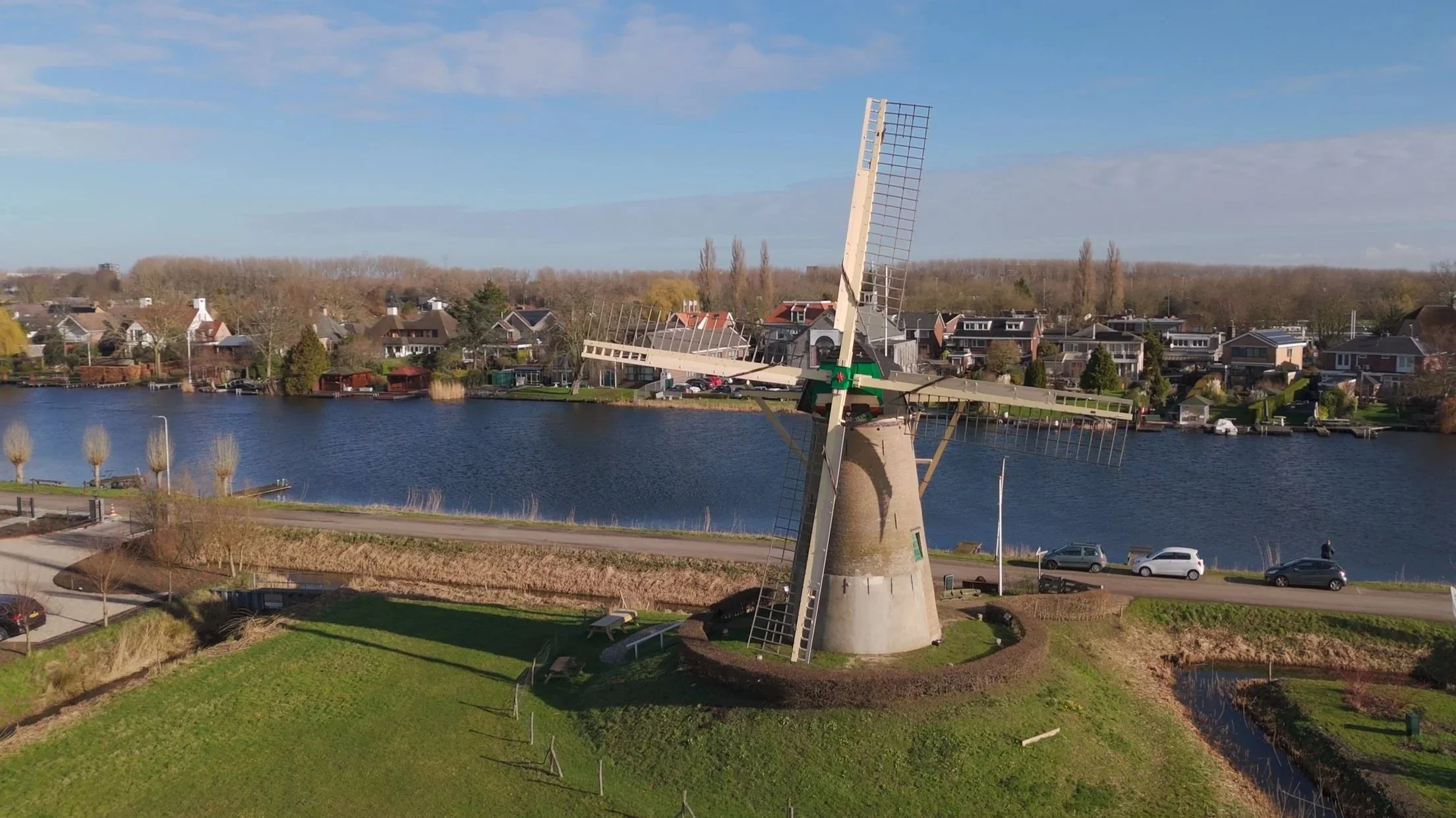 Een windmolen met vier wieken langs een rivier met huizen op de achtergrond onder een blauwe hemel.