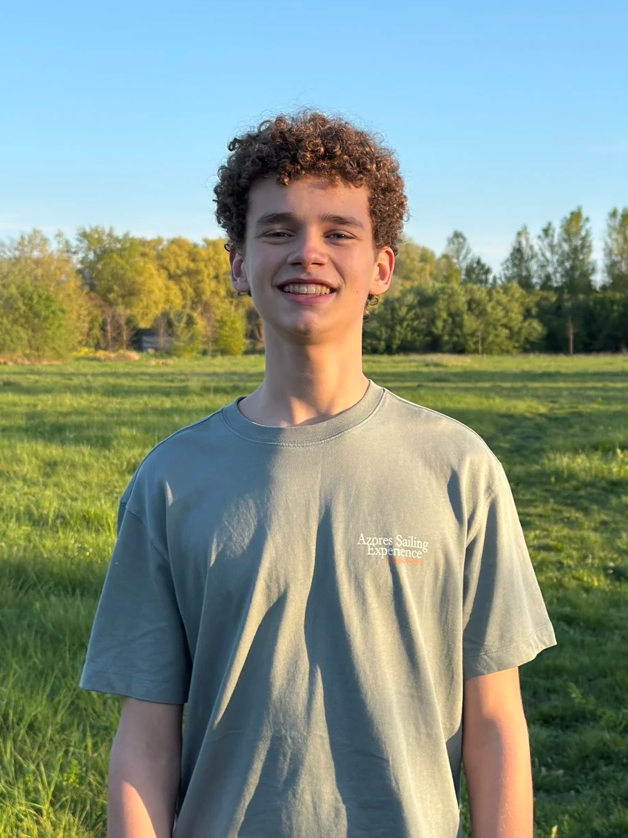 Een jonge man met curly haar staat buiten in een groen veld, lachend, met bomen en een heldere blauwe lucht op de achtergrond.