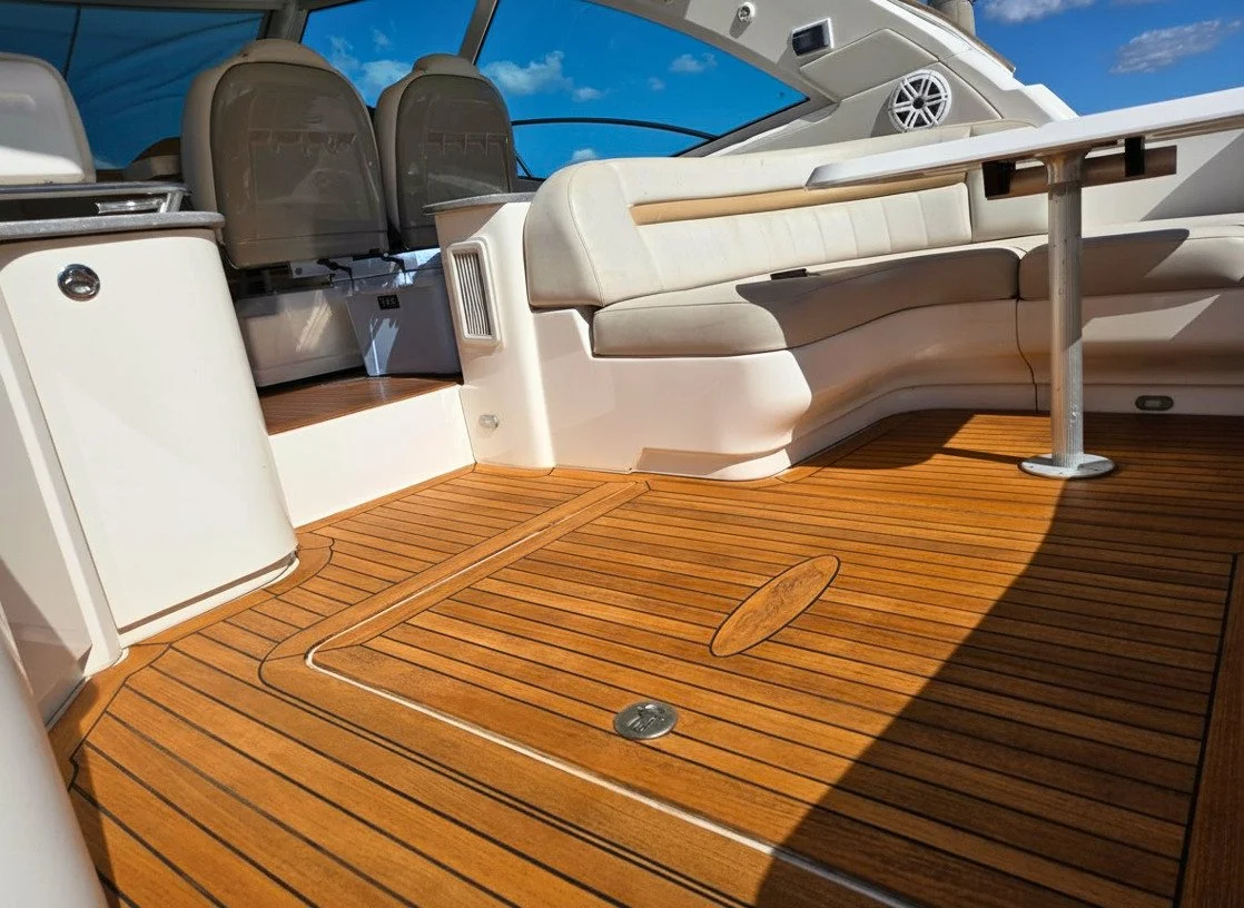 View of the deck area on a yacht, featuring tan cushioned seating, a teak floor, and a table with a metal post, with a clear blue sky in the background.