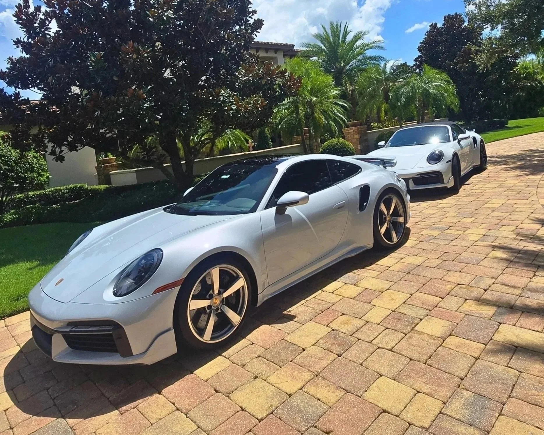 Two silver luxury Porsches parked on a brick driveway in front of a lush, landscaped yard with palm trees and a house in the background.