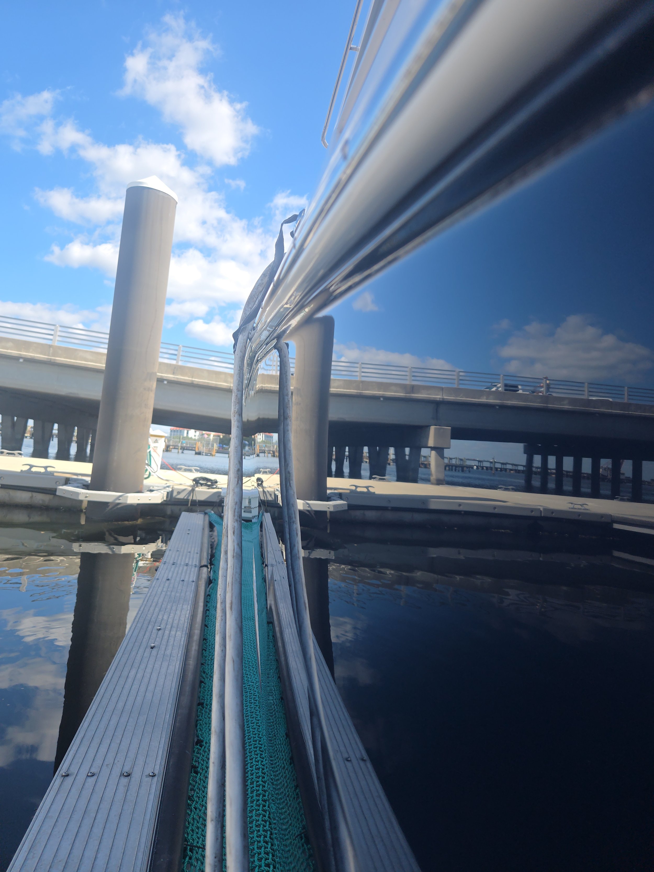 View from the dock of a boat with the boat's shiny metallic side reflecting the sky and clouds overhead, and the dock structure visible in the background.