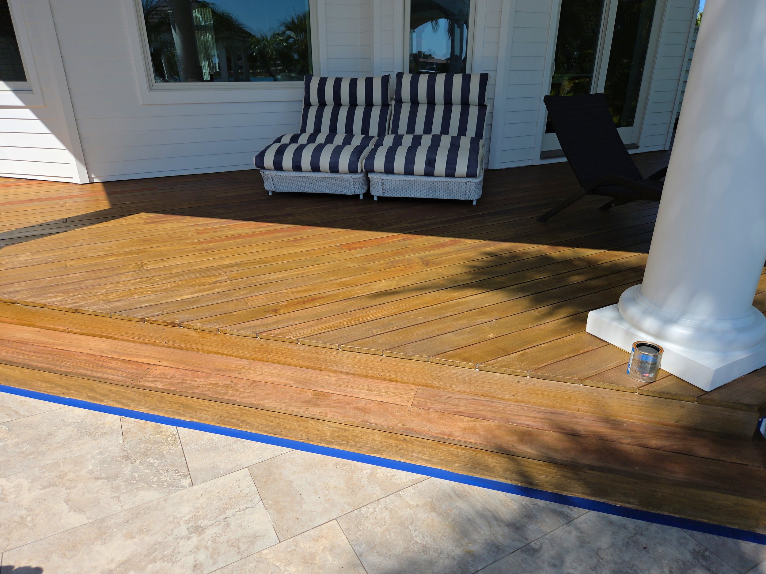 Luxury exotic wood deck attached to a house with white siding, patio furniture with striped cushions, a black lounge chair, and a white column with paint can nearby.