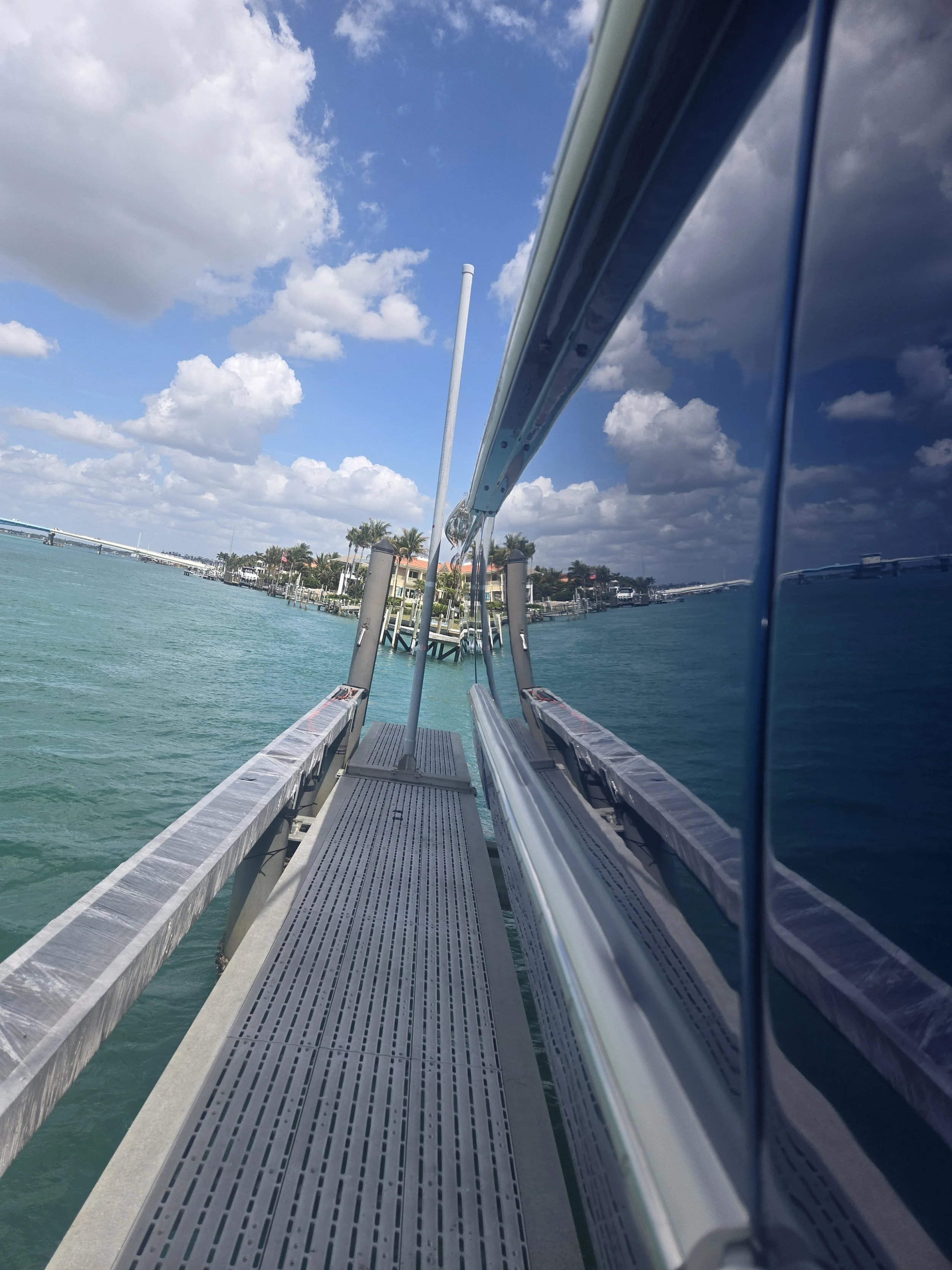 View from a boat dock showing a narrow metal walkway, a reflection of the boat's side on the boat's window, water, a marina with docks, houses, and palm trees under a partly cloudy sky.