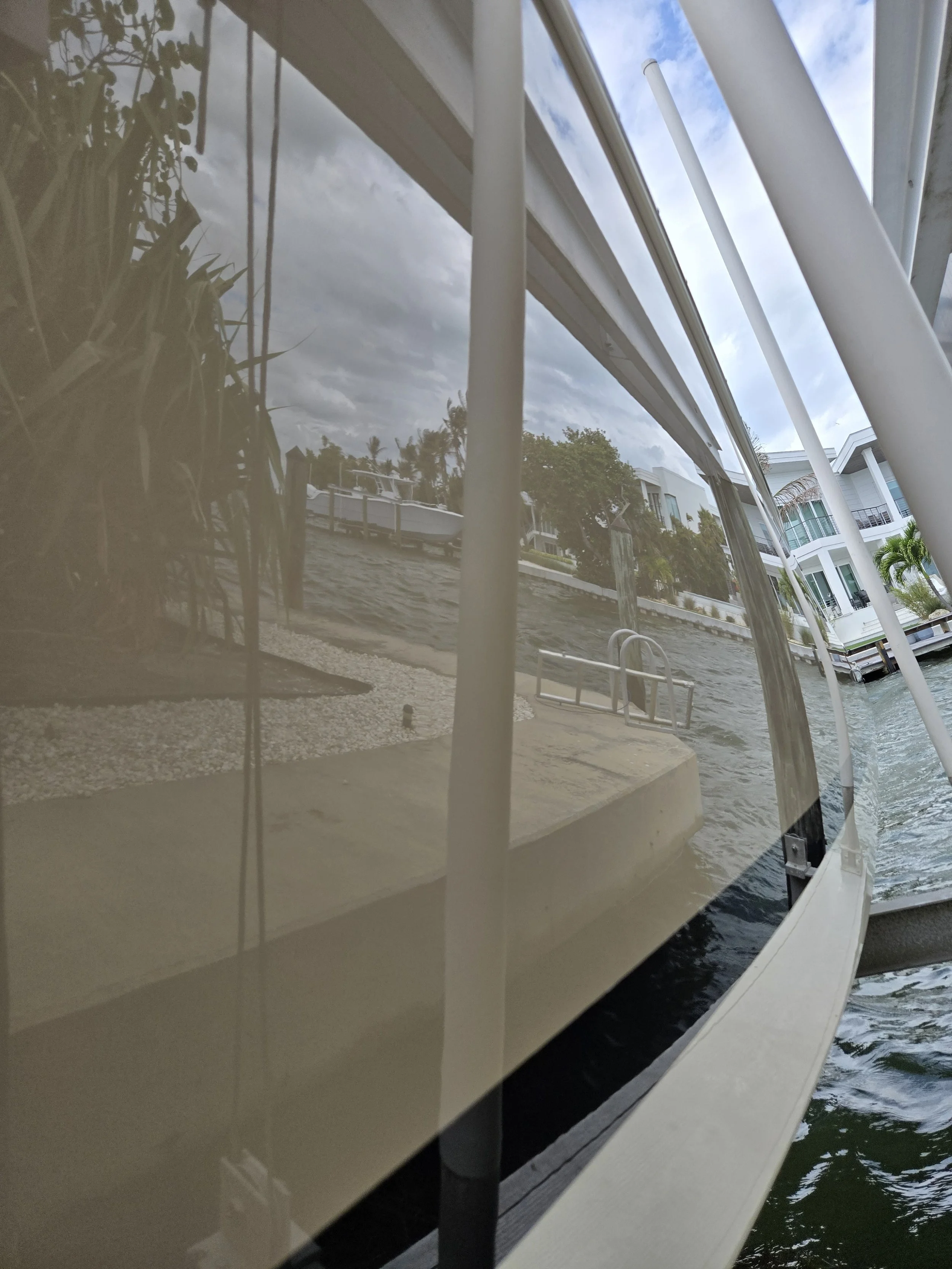 View of a dock with stairs and a Chris Craft, reflected in a glass window, with houses and trees along a waterway under cloudy skies.