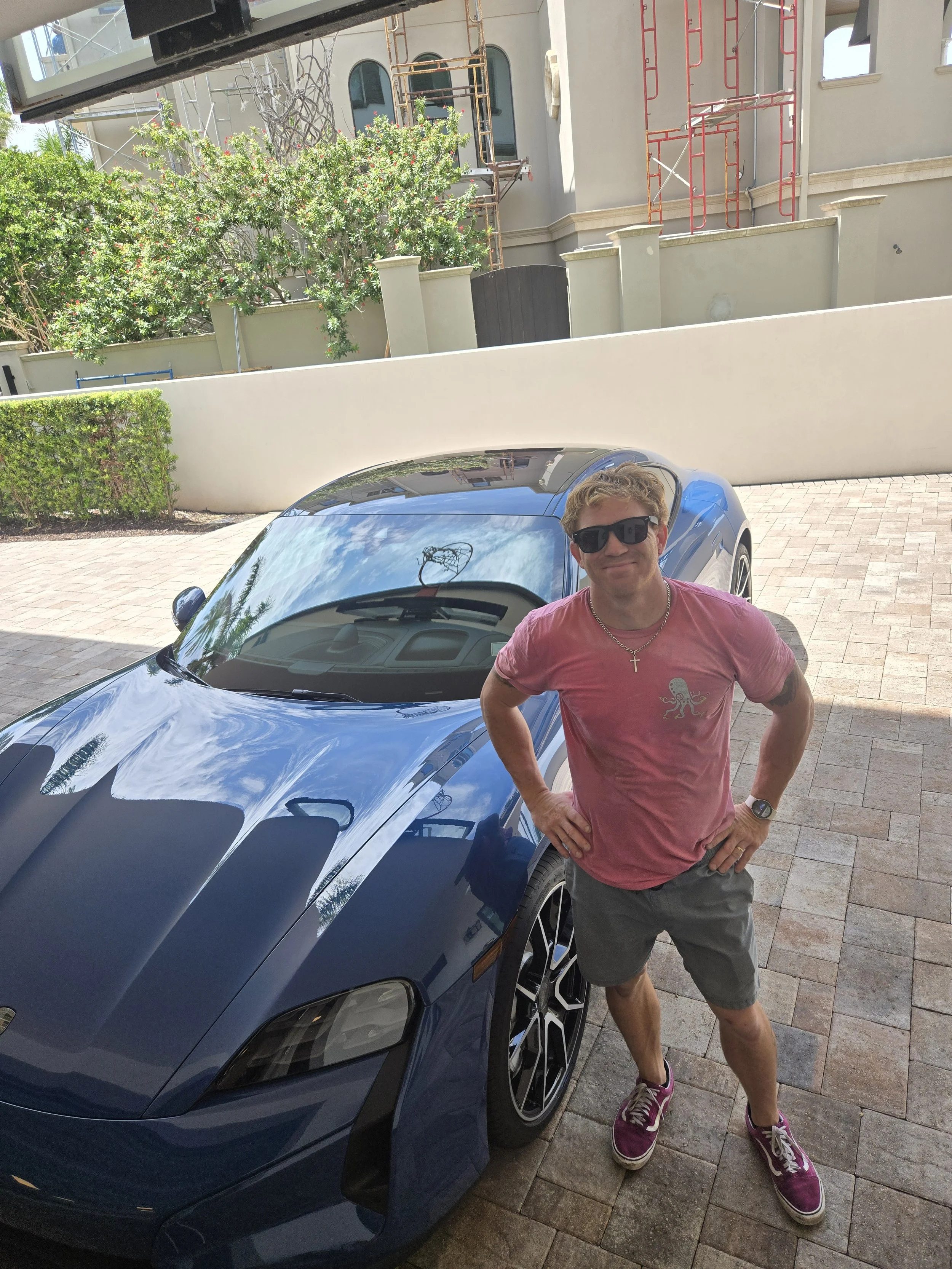 A man wearing sunglasses, a pink t-shirt, gray shorts, and purple sneakers stands confidently with hands on hips beside a blue electric Porsche in a driveway with a building and greenery in the background.