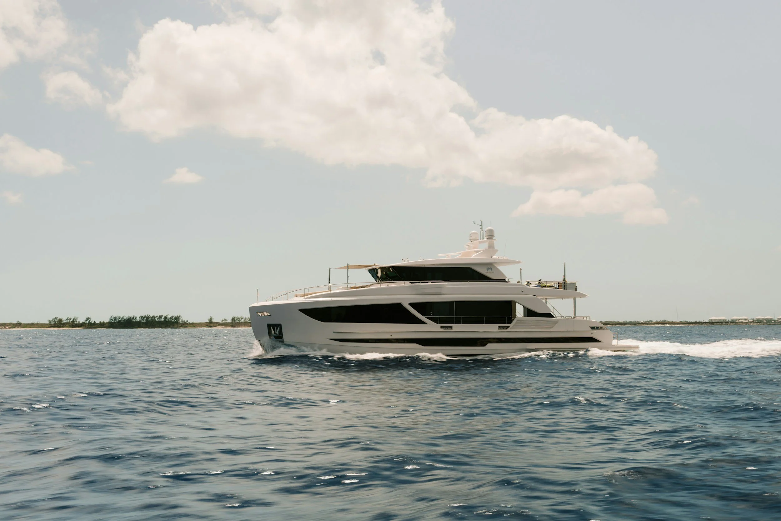 A large white yacht cruising on the water with a distant shoreline and trees, under a partly cloudy sky.