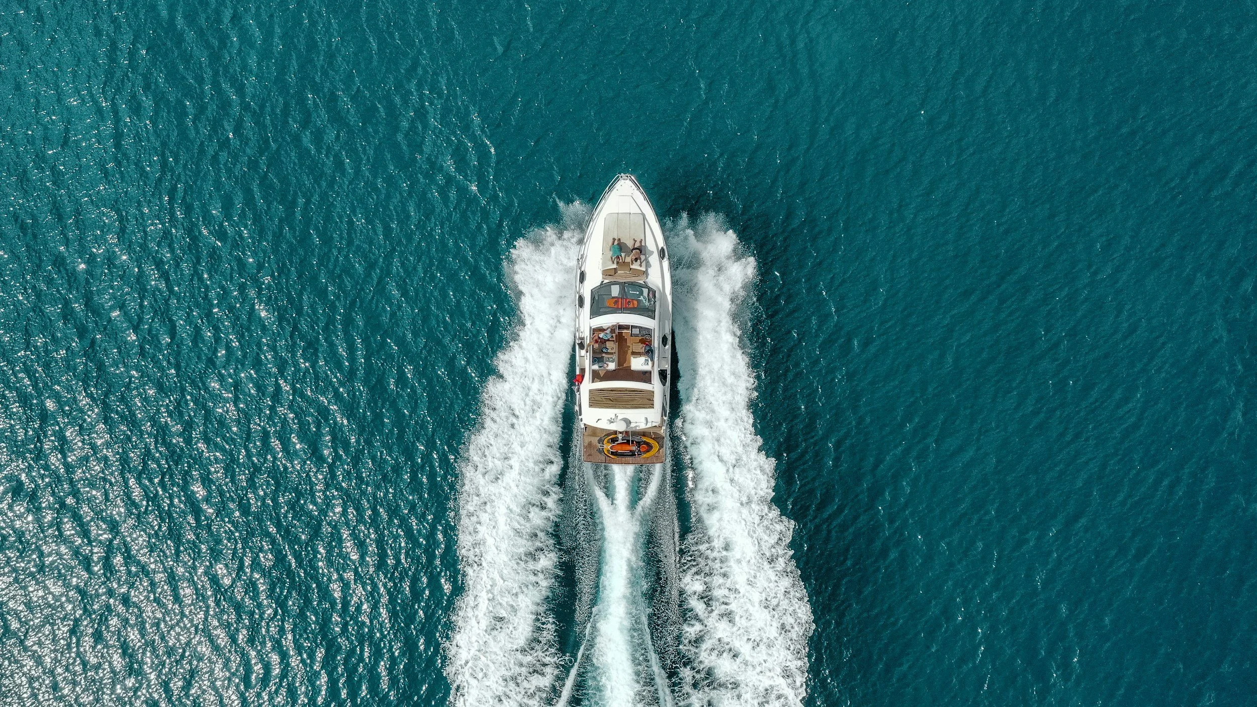 An aerial view of a motorboat speeding across the ocean, creating white wake, with two people on board relaxing on the deck.