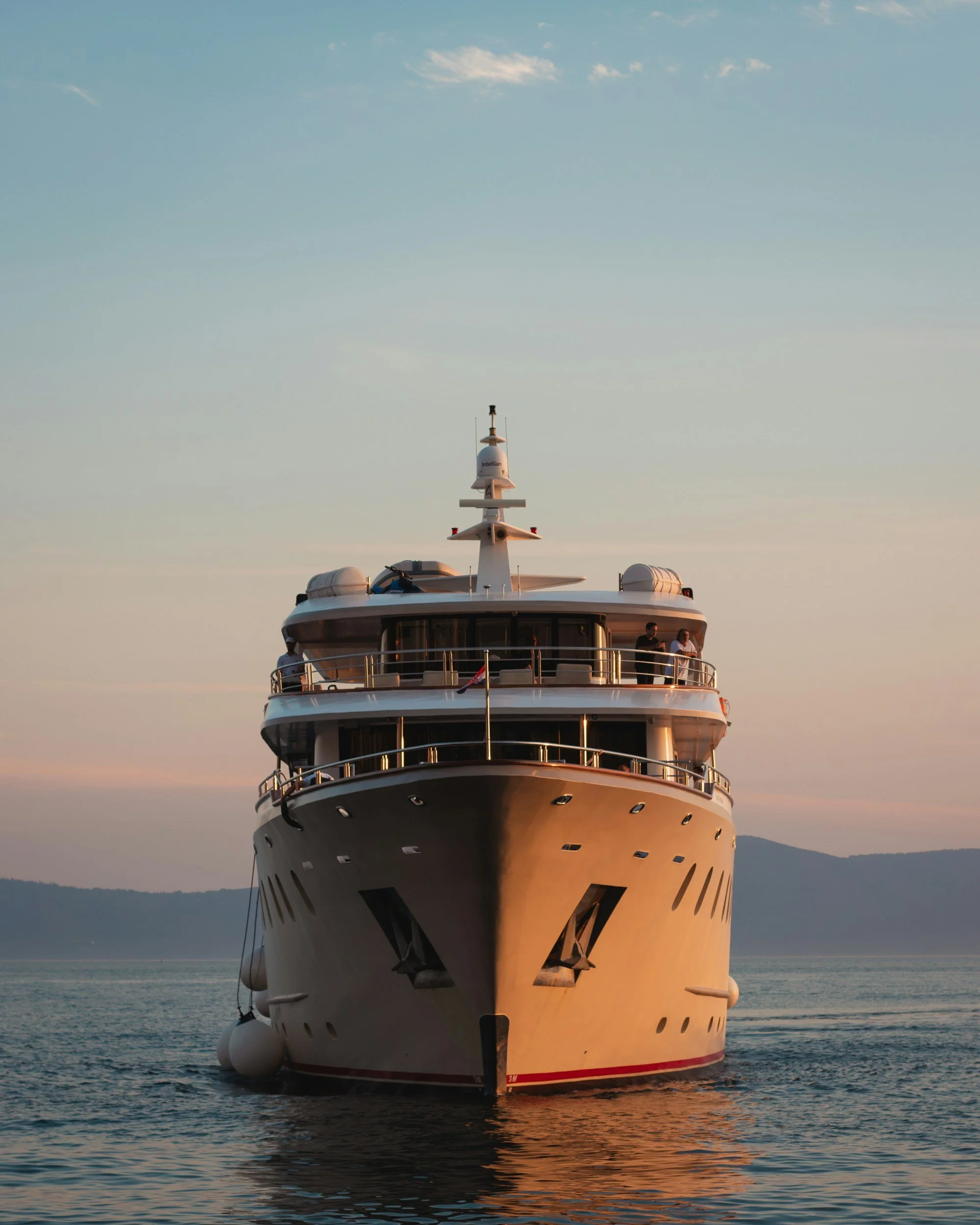 Front view of a large luxury yacht sailing on the ocean during sunset with a mountain in the background.