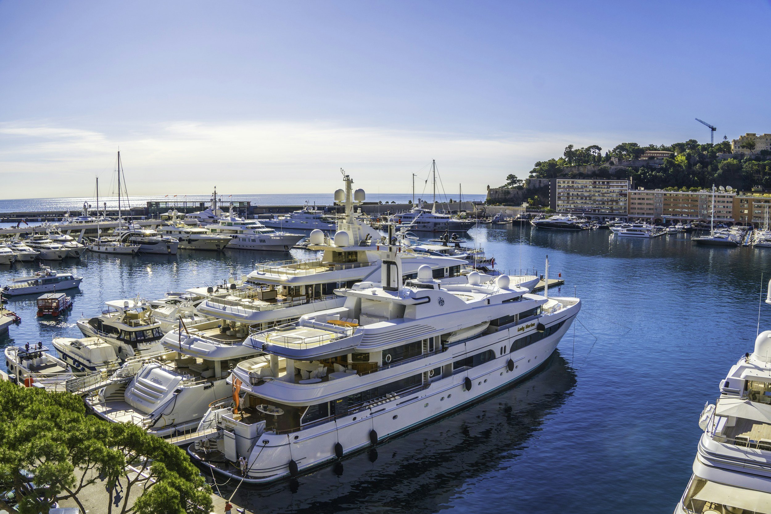 A marina filled with luxury yachts docked by a coastline with multi-story buildings and green hills in the background on a sunny day.