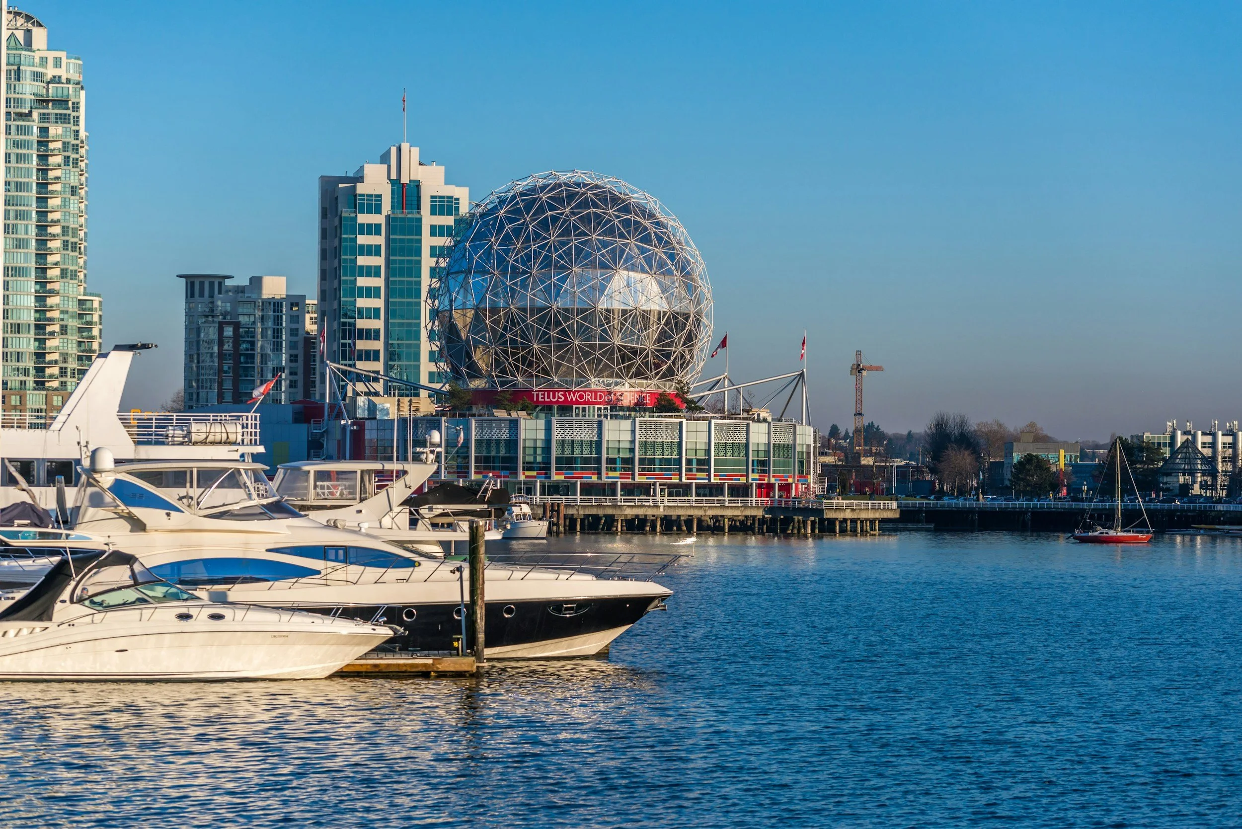 Marina with boats in the foreground and a city skyline including a geodesic dome and high-rise buildings in the background under a clear blue sky.