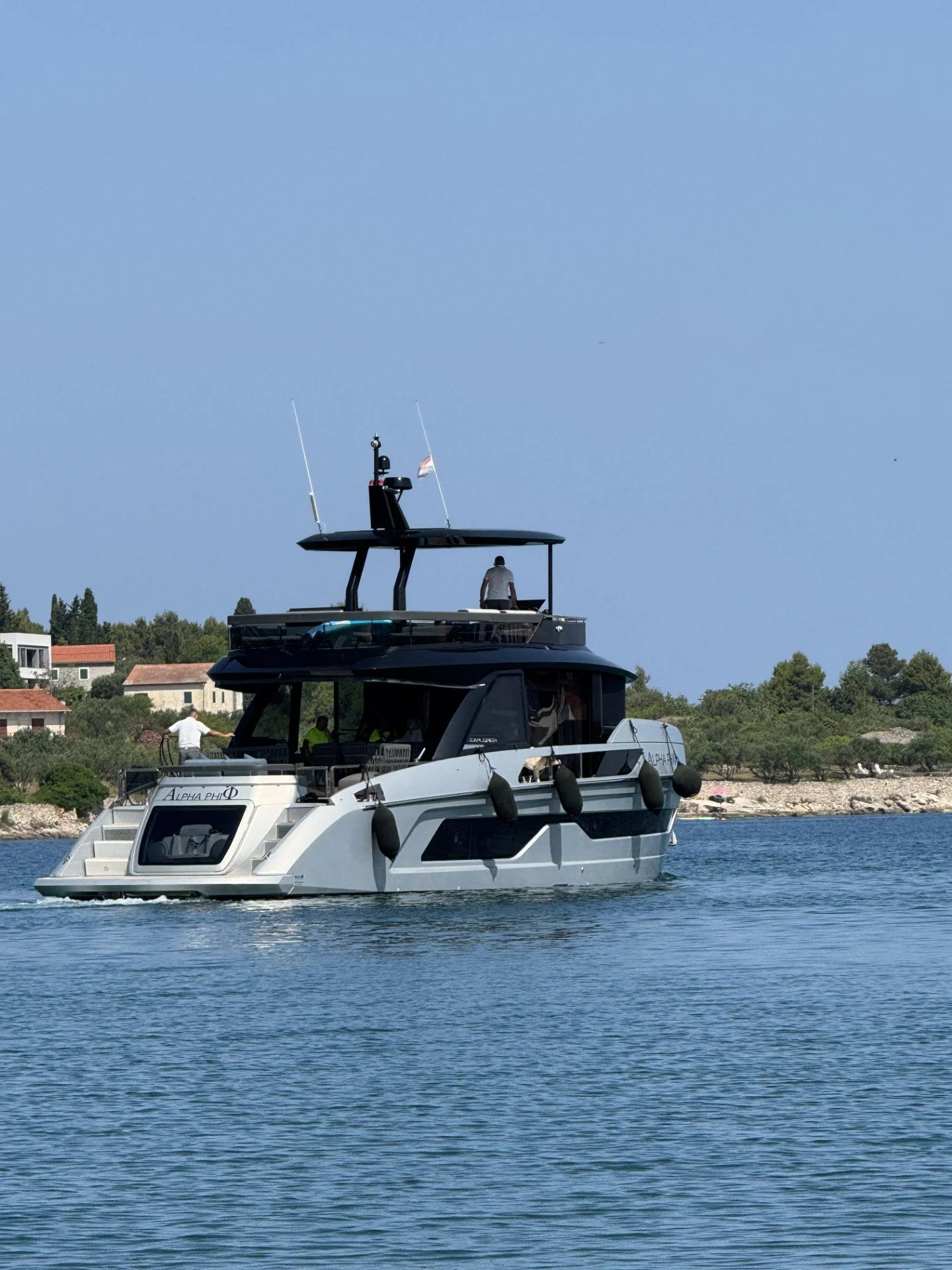 A luxury yacht named Alpha Phi sailing on calm water near a shoreline with houses and trees under a clear blue sky.