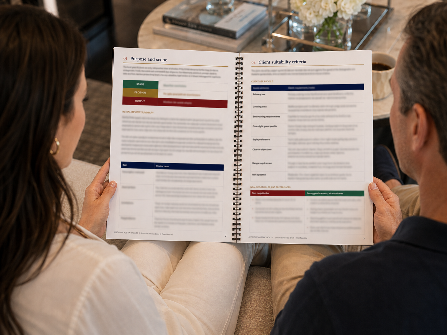 Two people sitting on a sofa, reading a spiral-bound document with charts and text, in a living room with a coffee table holding a glass vase of white flowers, a coffee mug, and books.