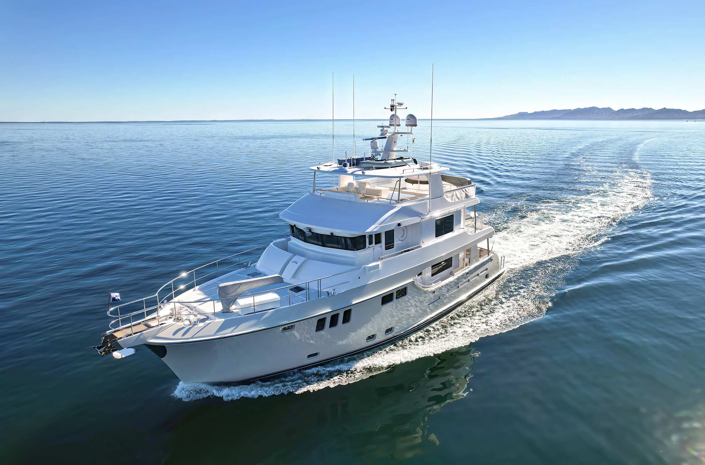 A white yacht sailing on calm blue water, with a distant mountain range in the background under a clear sky.