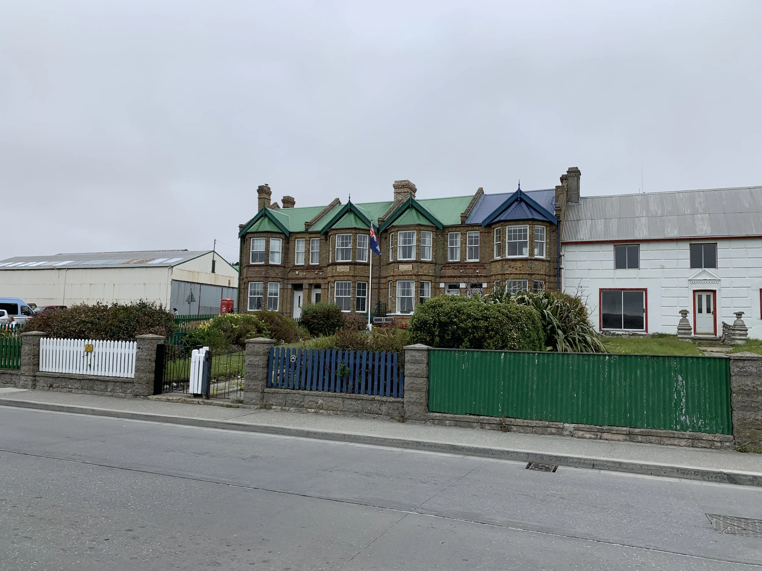A row of buildings along a street, including a historic brick building with green and blue rooftops, a white building with a red door and window, and various shrubs and plants in the foreground under cloudy skies.