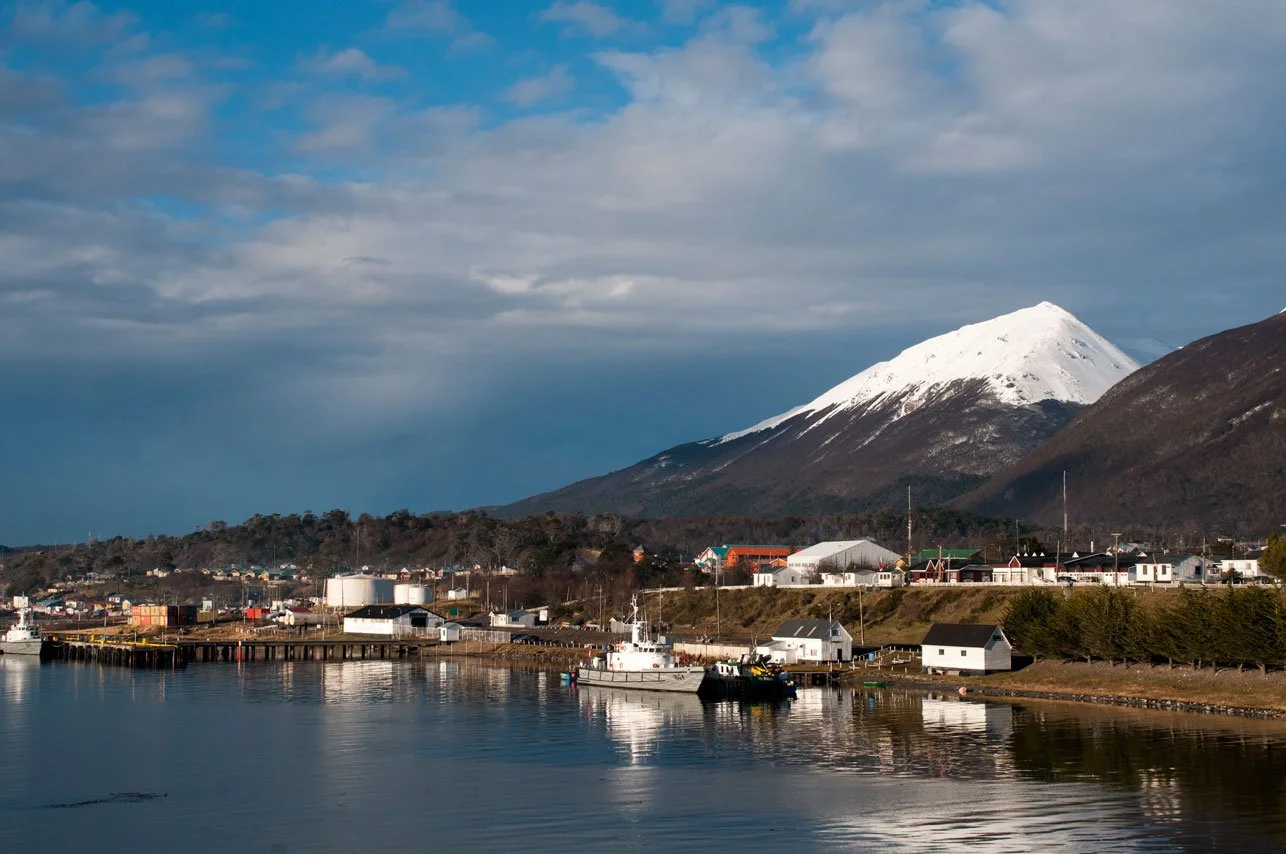 A coastal town with a docked boat, small houses, and industrial storage tanks, with a snow-capped mountain and cloudy sky in the background.
