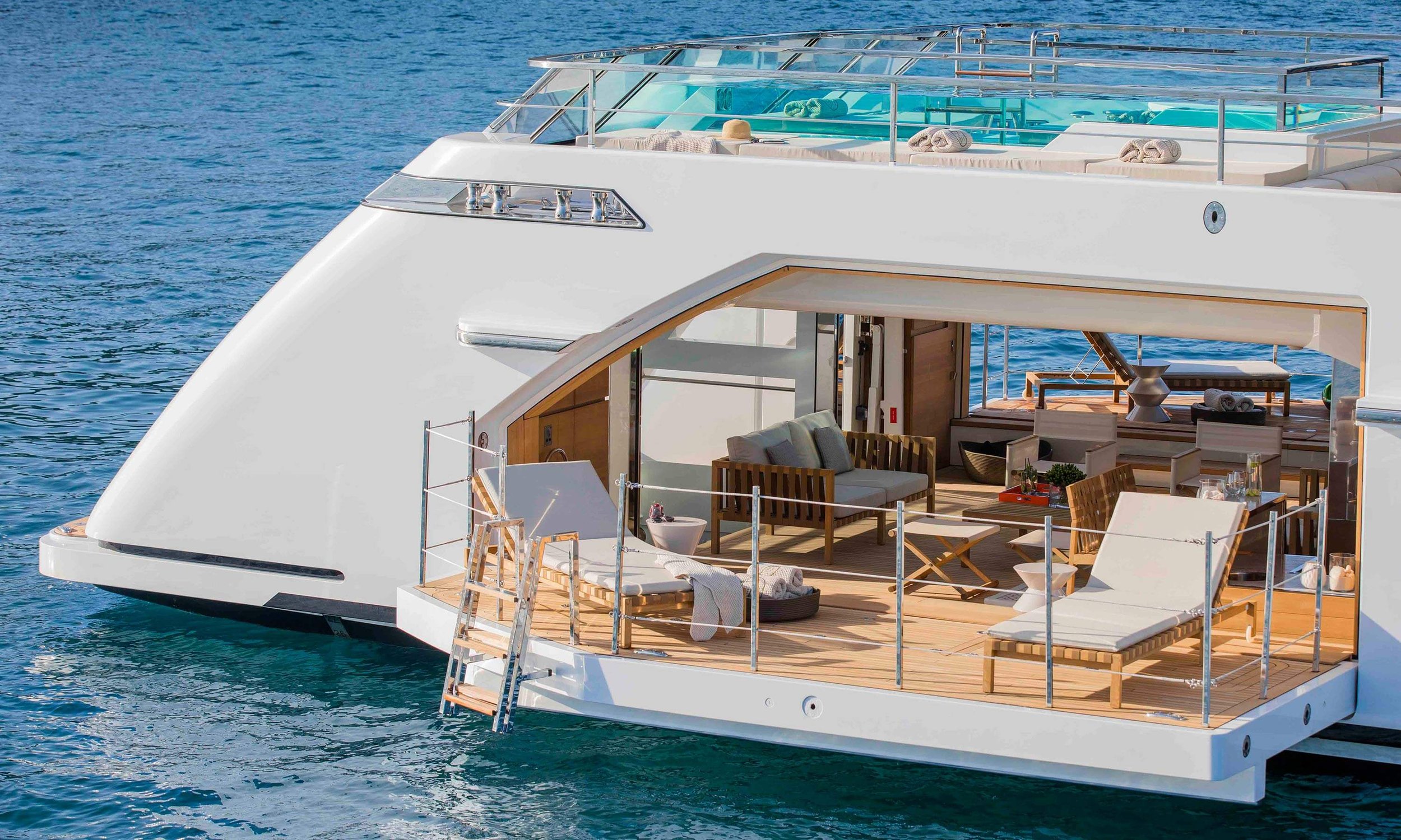 A luxury yacht floating on the water with an outdoor seating area, sunbeds, towels, and a pool area on the upper deck.