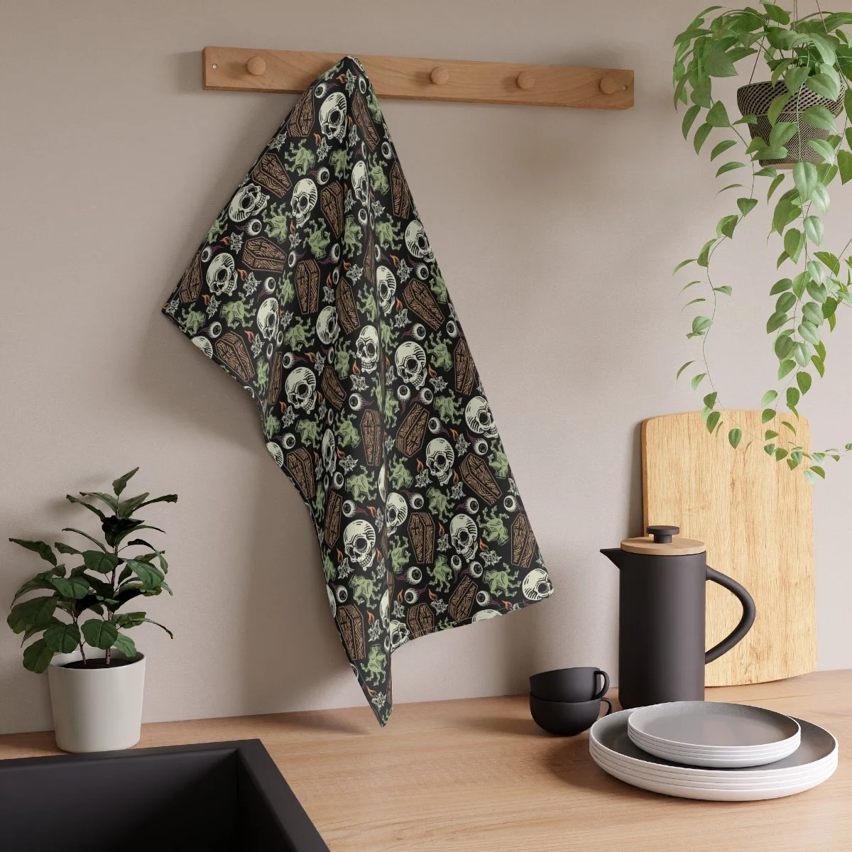 Kitchen with a wooden countertop, black and white plates, a black thermos, a small black cup, a wooden cutting board, and hanging cloth with a skull and coffin pattern, surrounded by plants.