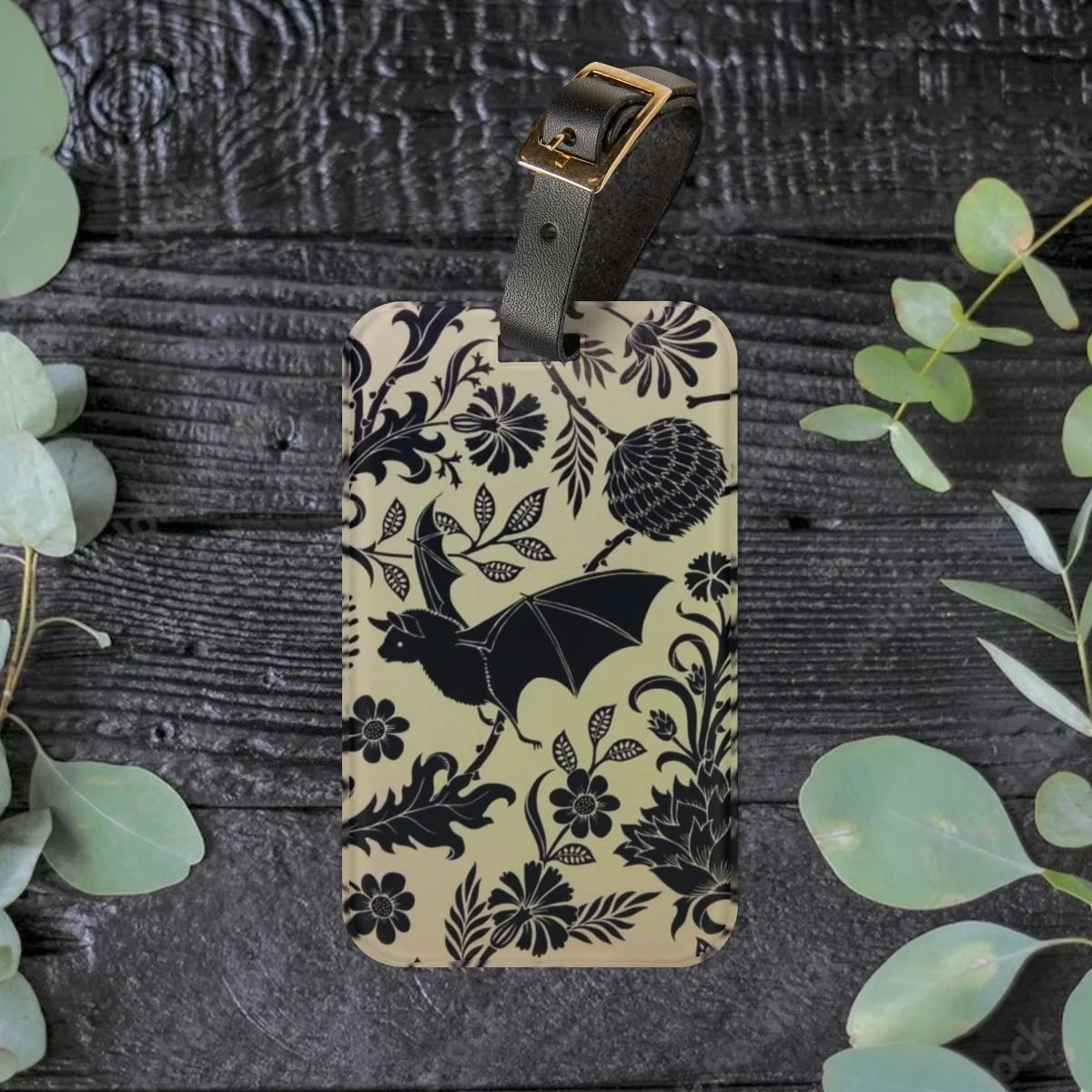 A luggage tag with a black floral and bat illustration on a beige background, placed on a dark wooden surface with eucalyptus leaves around it.