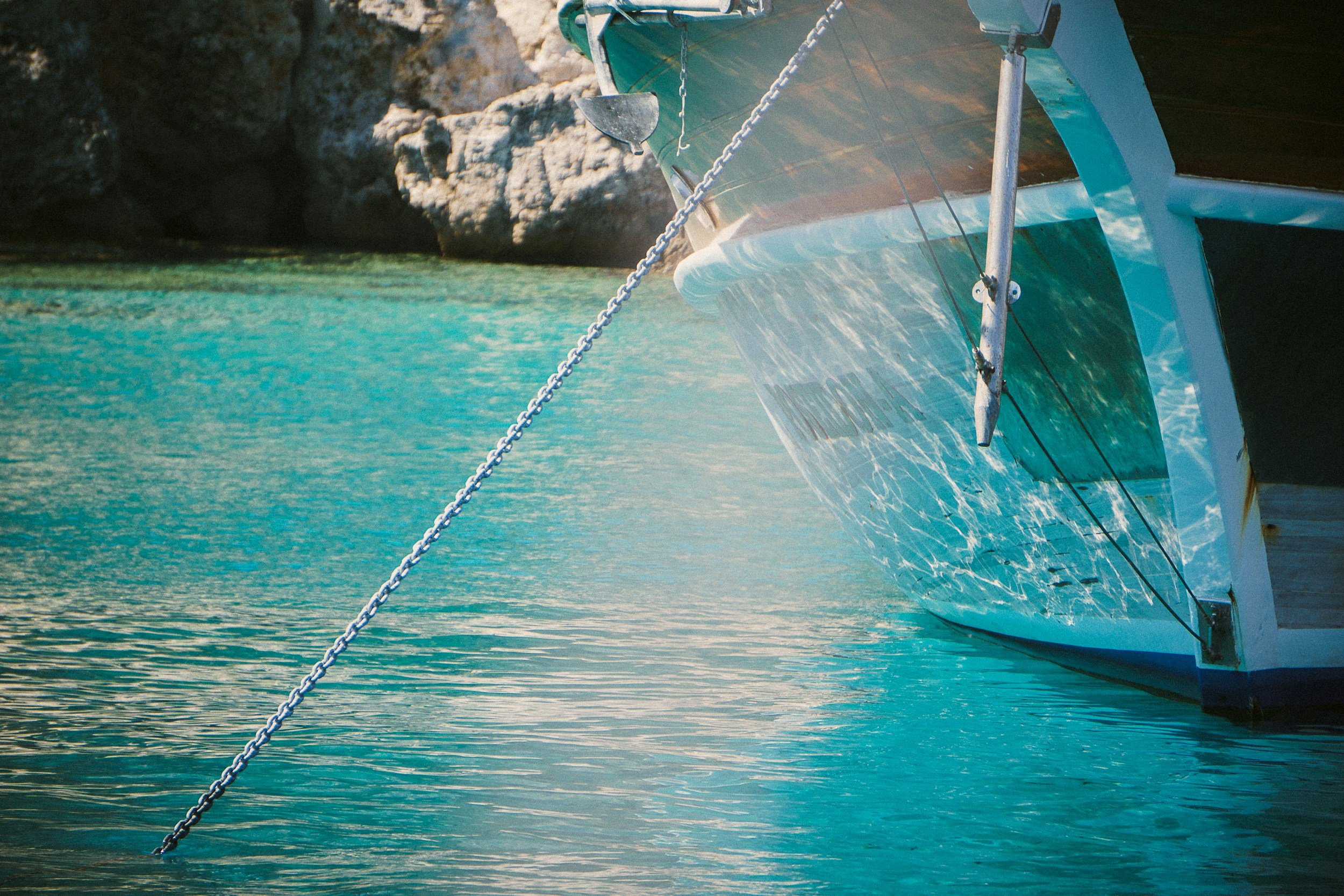The front of a boat docked in turquoise water near rocky cliffs, with a chain securing the boat.