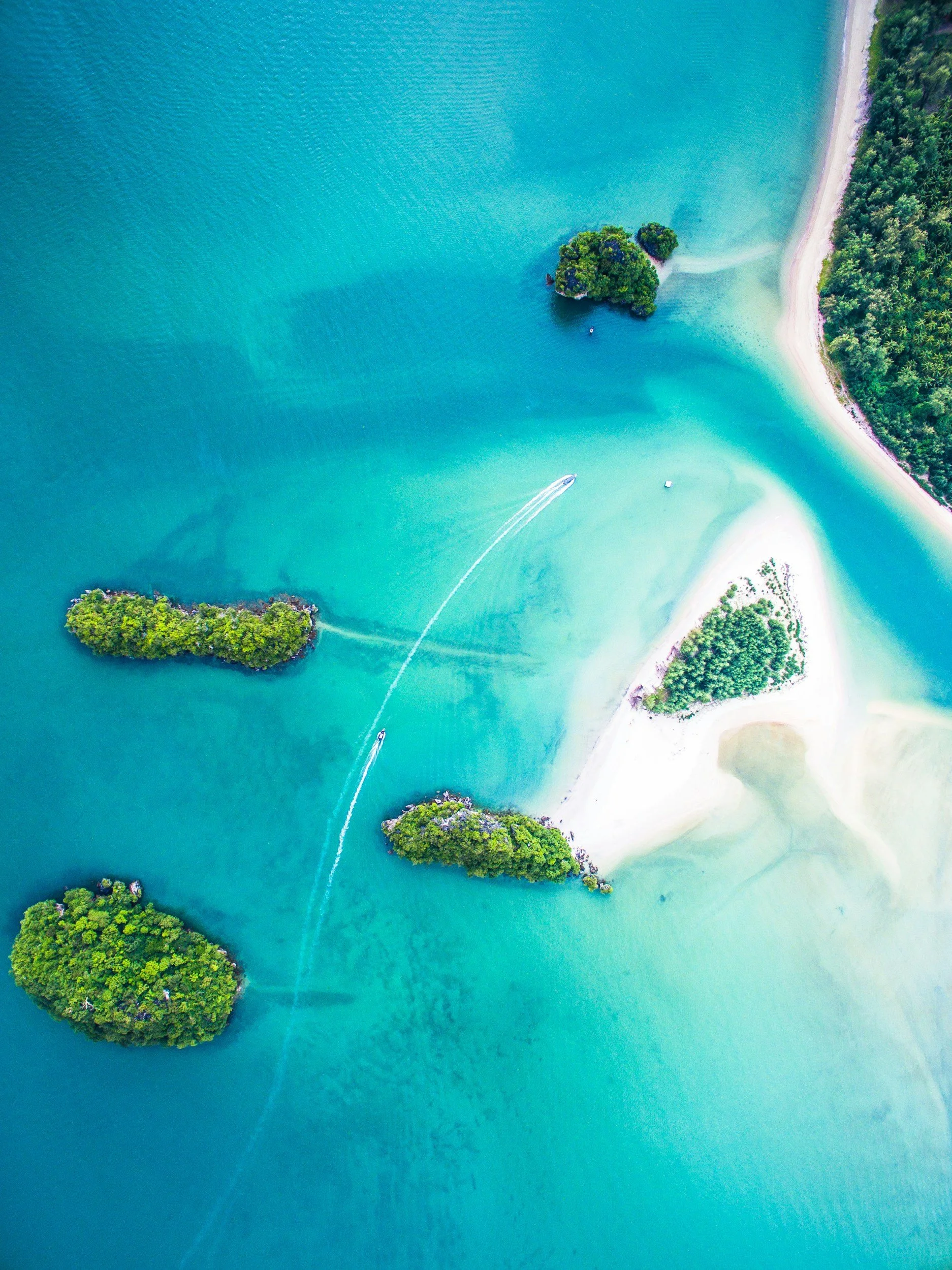 An aerial view of small lush green islands surrounded by bright turquoise water near a sandy beach with some greenery.